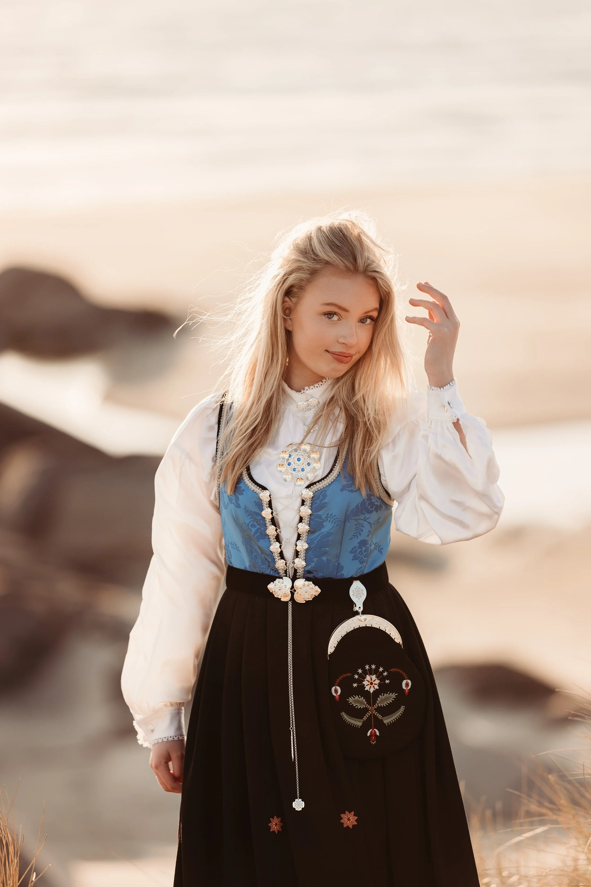 En ungdomspike i tradisjonell norsk bunad står på stranden ved sjøen under solnedgang, ser mot kamera og smiler.