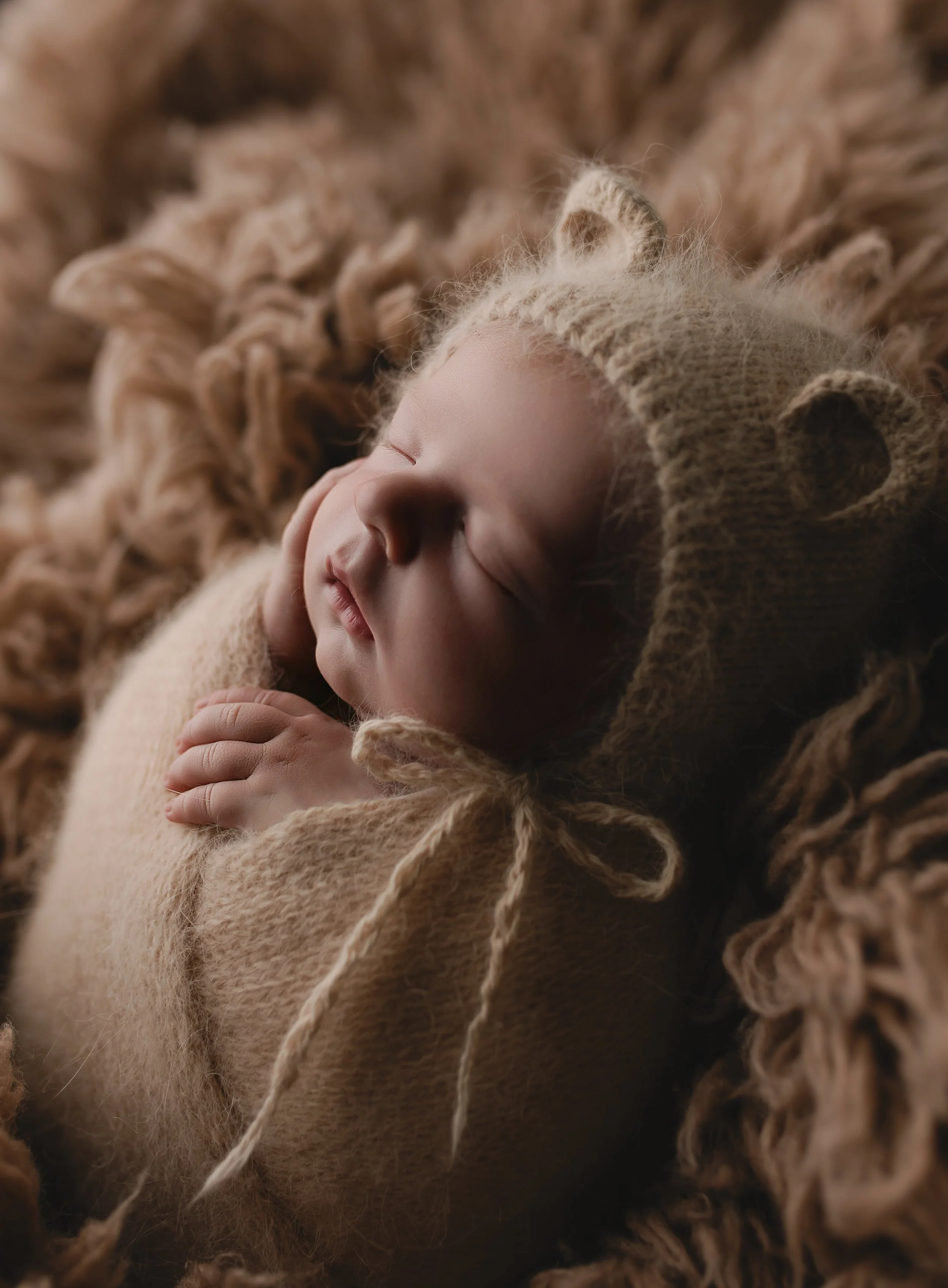En sovende baby kledd i en beige kofte med en matchende lue med bamseører, liggende på en myk, sherpa-lignende overflate.
