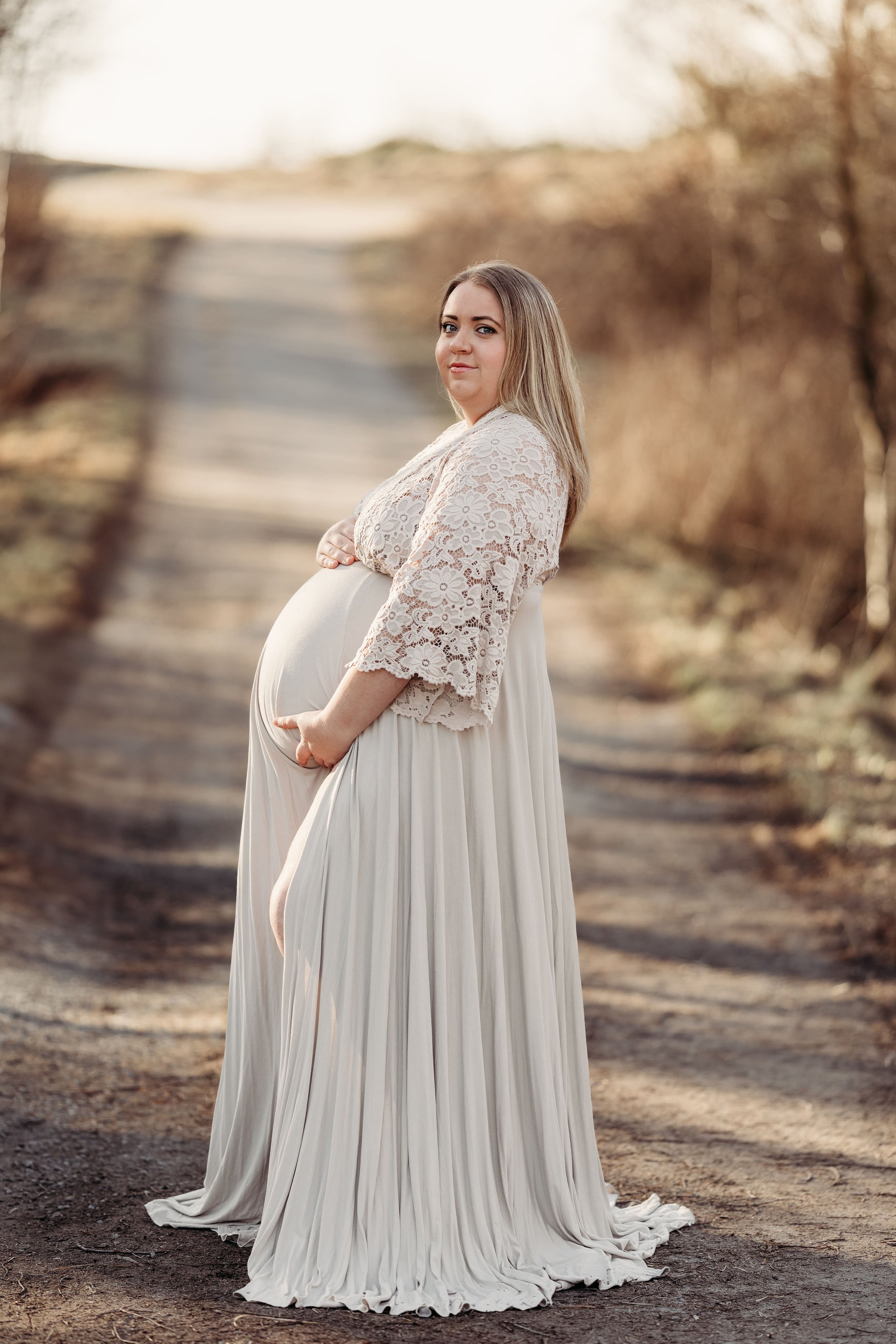 En gravid kvinne står på en jordvei i solnedgangen, iført en lang, hvit, plyset kjole og en blondejakke.