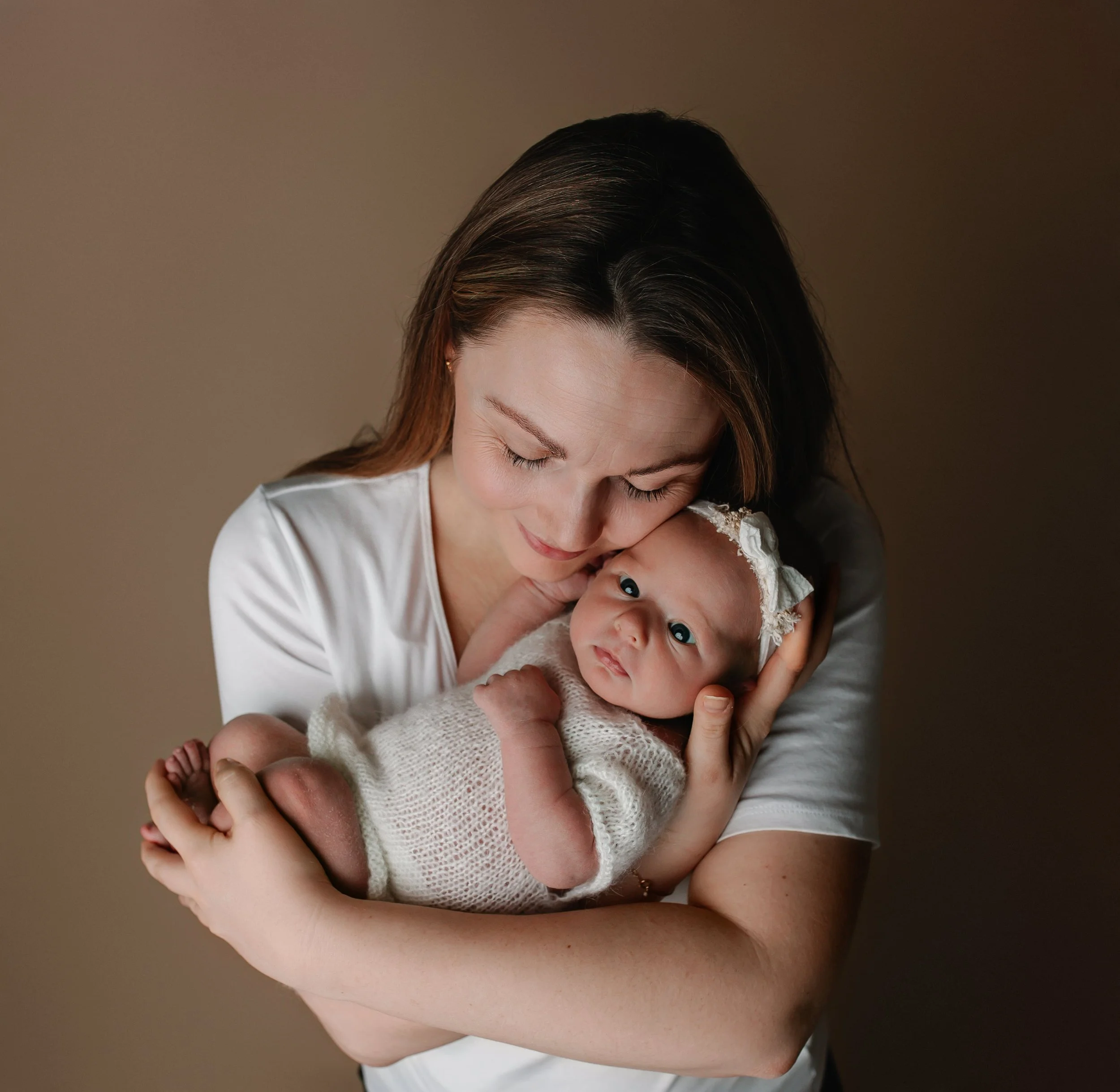 Et kvinne som klemmer en baby, som har på seg en hvit, strikket-stau og et hvitt hodebånd med blonder. Begge har lyse hudtoner og kvinnen har brunt hår.