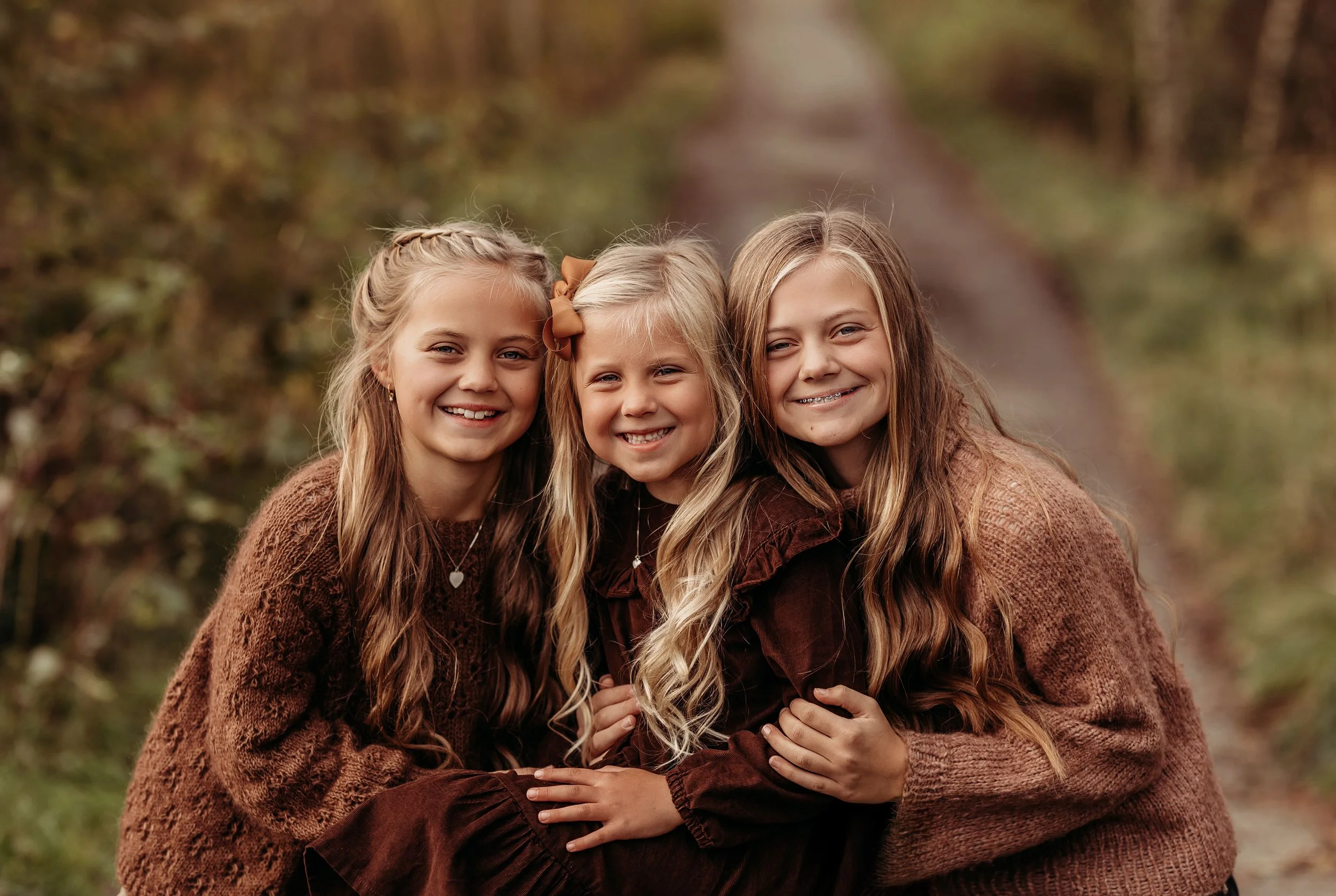 Tre unge jenter som smiler og klemmer hverandre utendørs i høstlandskap.