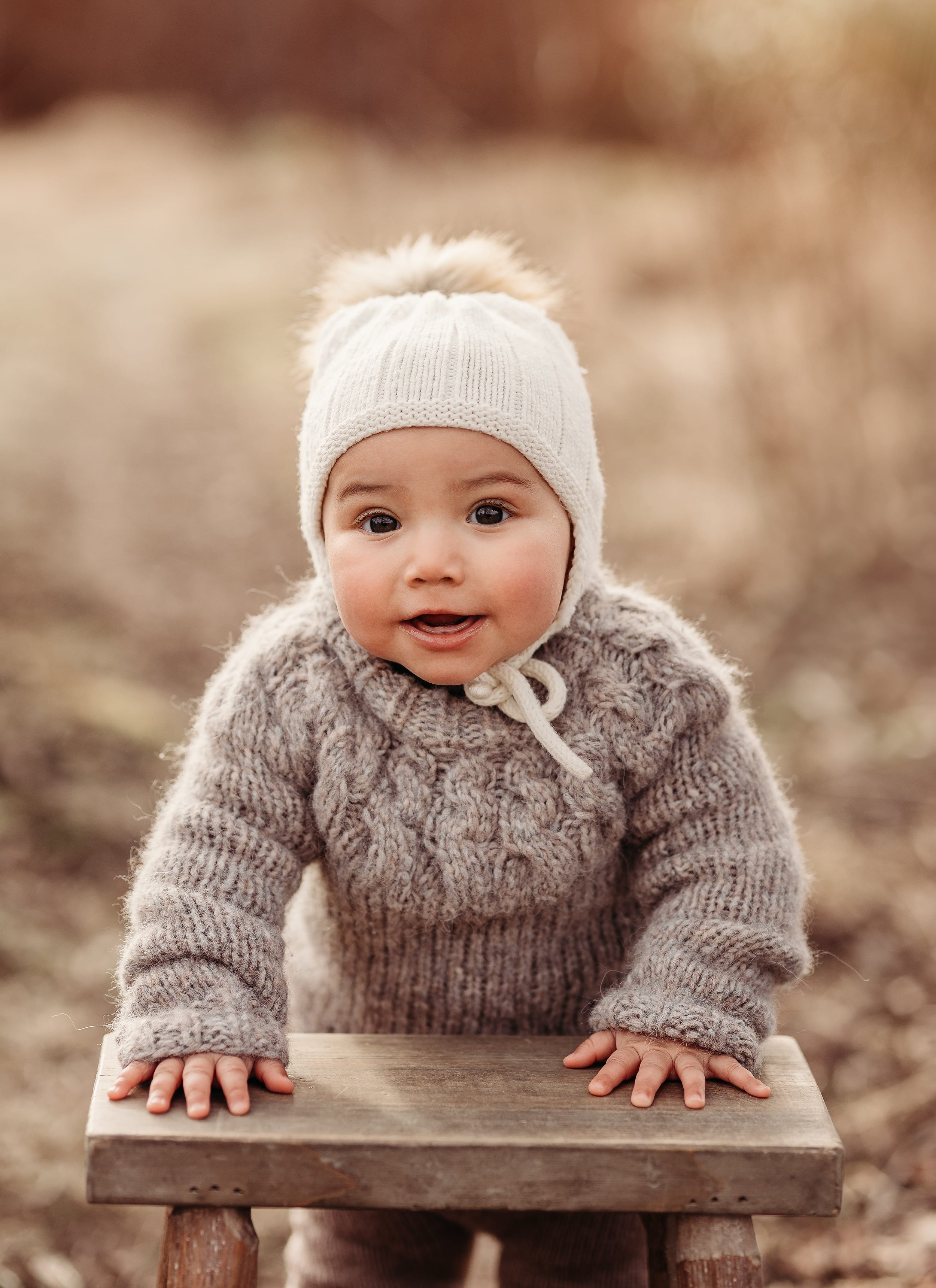 Et lite barn med påfuglfarget genser og hvit lue som står med hendene på en skjenk, ute i et område med høstløv og naturlig belysning.