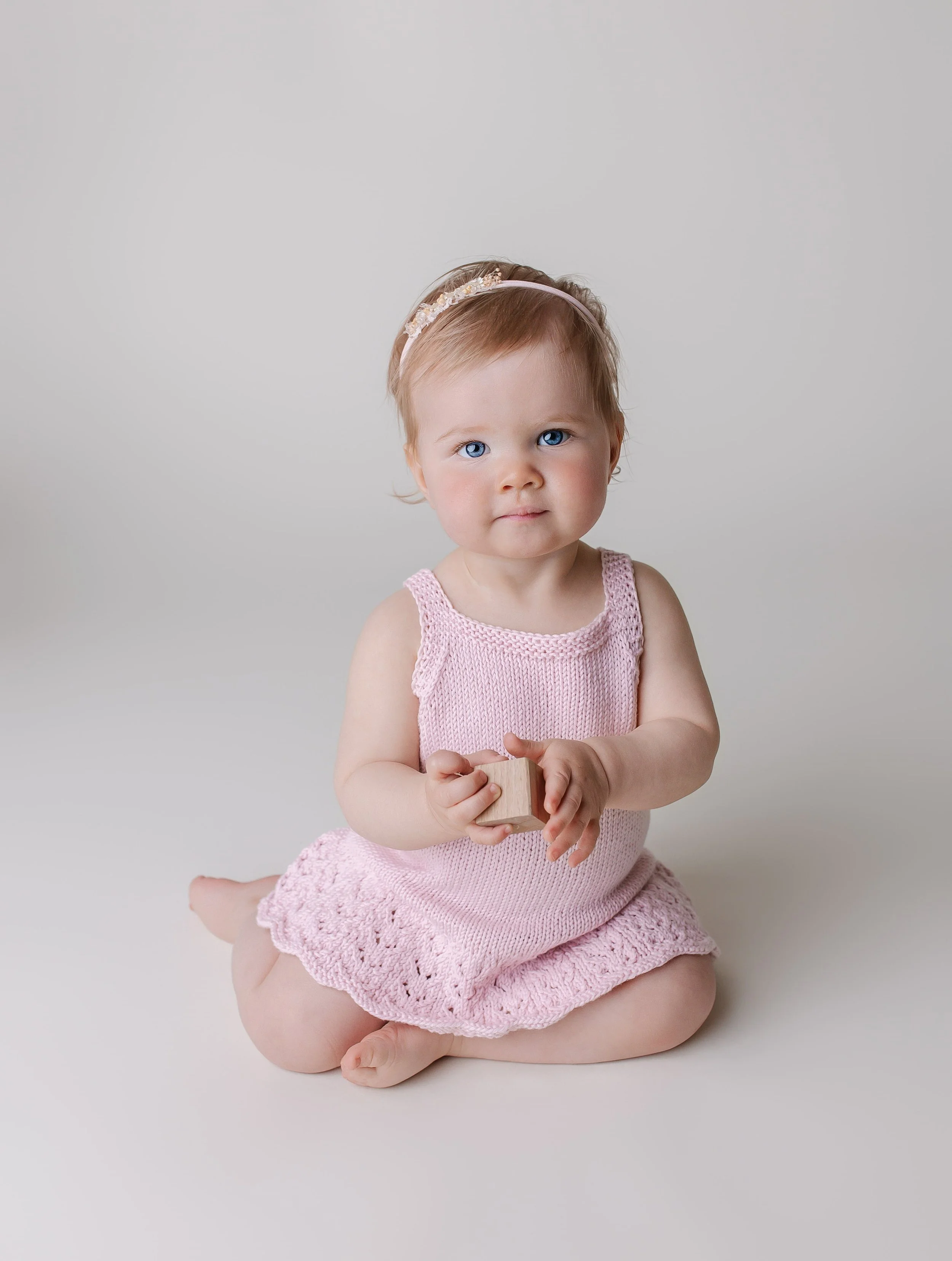 Liten jente med blond hår og blå øyne, iført rosa heklet kjole med blondekanter, sitter på gulvet med en liten trekloss i hendene, foran en lys grå bakgrunn.