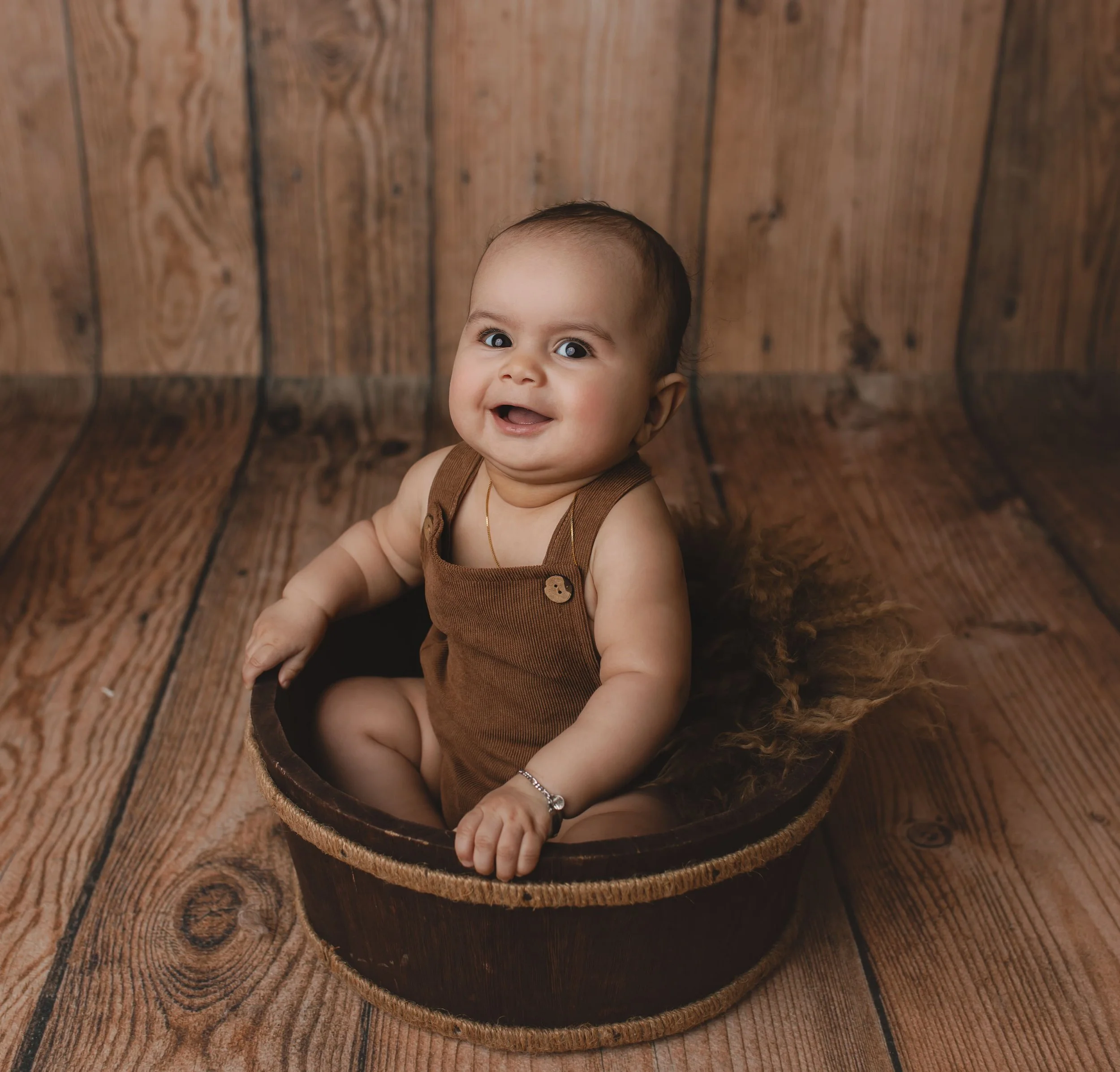 Smilende baby i brunt antrekk sittende i en rund kurv på tregulv med trefargede vegger i bakgrunnen.