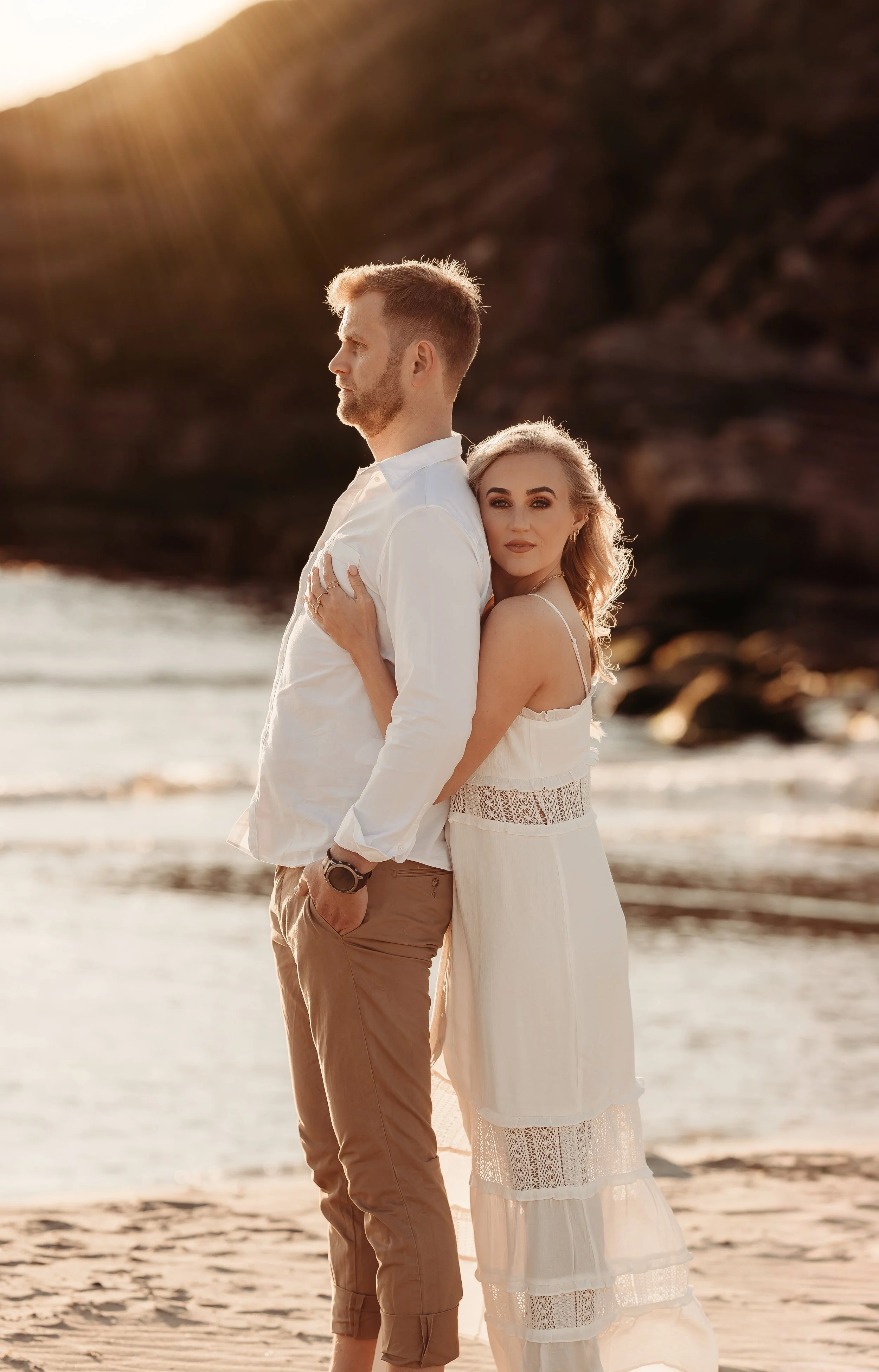 Et par står på stranden ved sjøen under solnedgang. Mannen har på seg hvit skjorte og brunt bukser, og kvinnen har på seg en hvit kjole. Kvinnen omfavner mannen bakfra, og begge ser rolig ut.