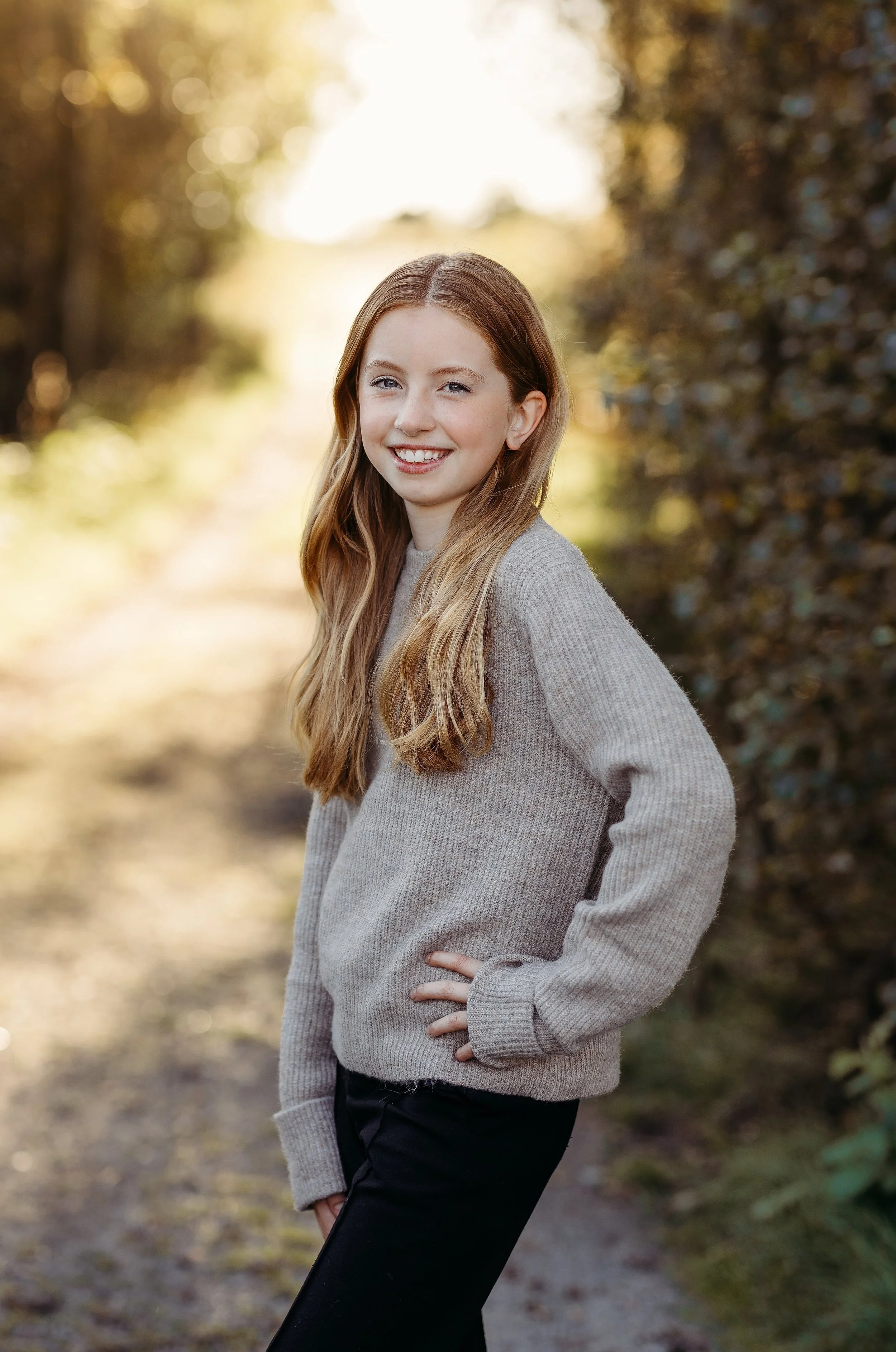 En smilende jente med langt, lyst hår, iført en grå genser og svarte bukser, står i en skogssti med høstfarger i bakgrunnen.