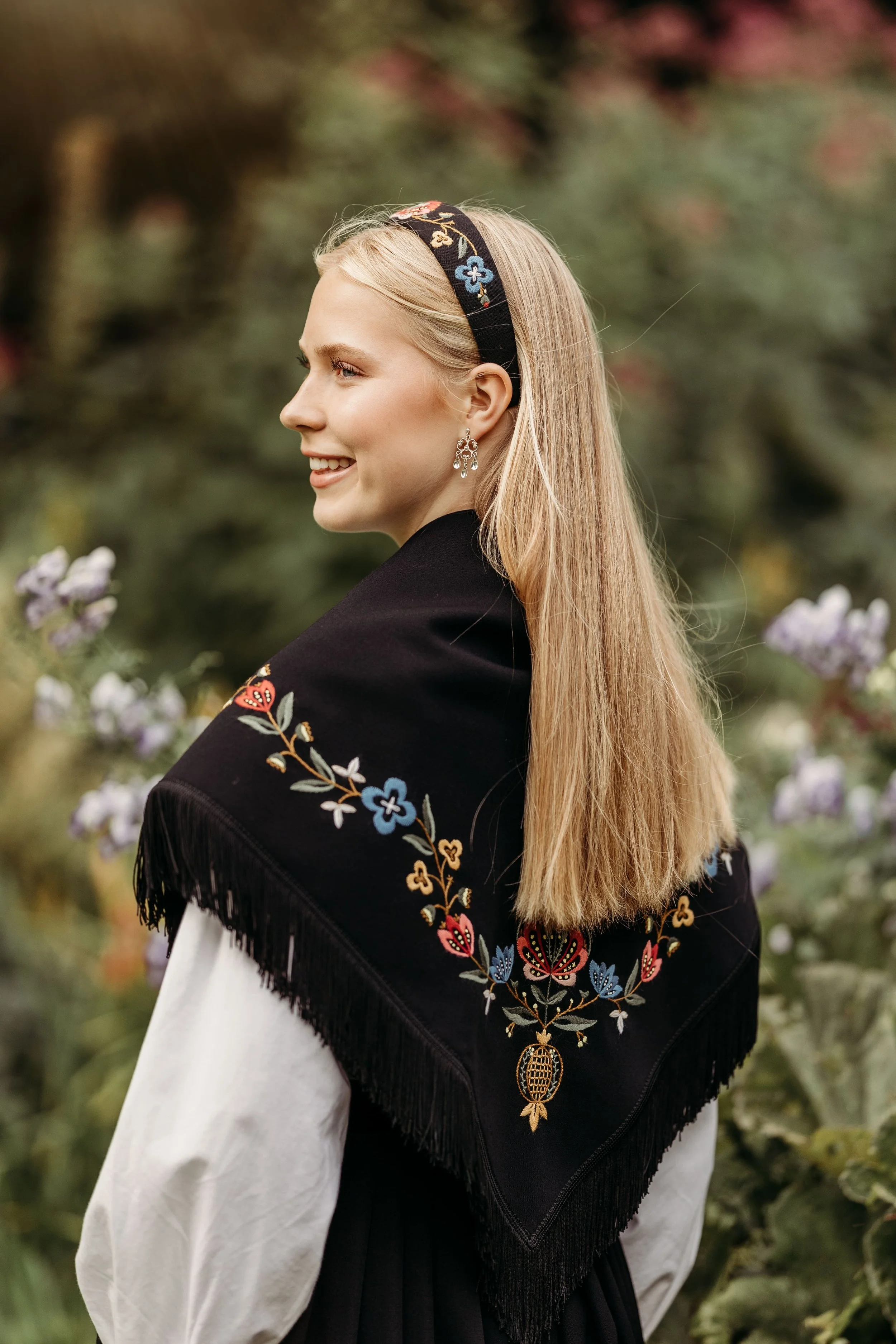 En kvinne med lang blond hår, smilende, har på seg en svart skjørt med blomsterbroderi og en svart brodert hodebånd, står utendørs blant blomster.