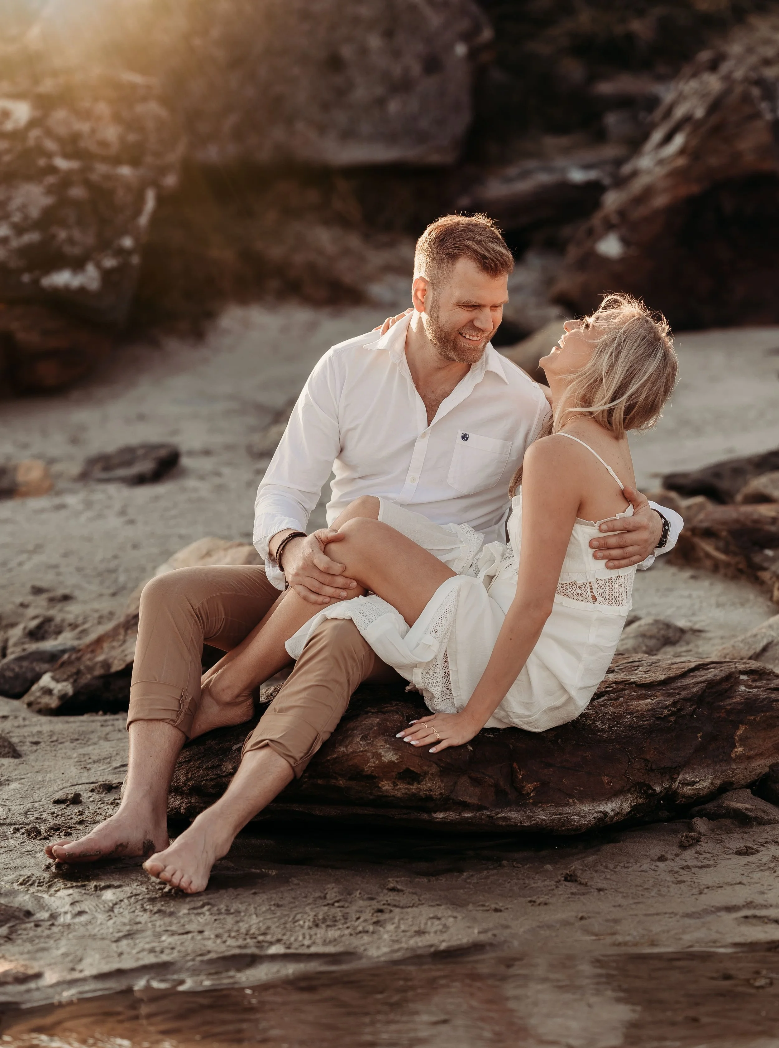 Et kjærlighetsfullt øyeblikk mellom et ungt par på stranden, omgitt av steiner og solskinn.