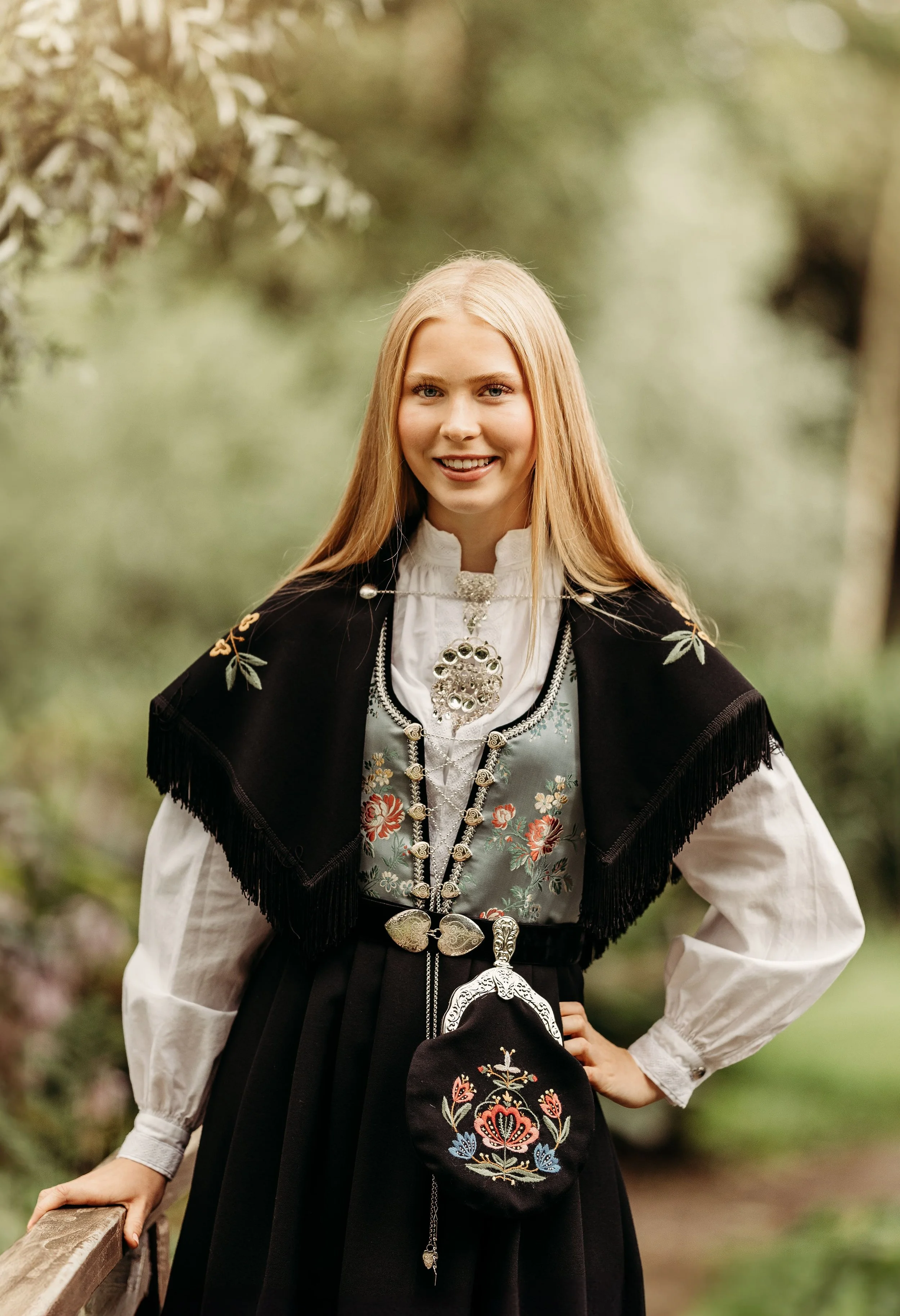 En ung kvinne i tradisjonell norsk bunad står ute i en grønn park, smilende mot kamera.