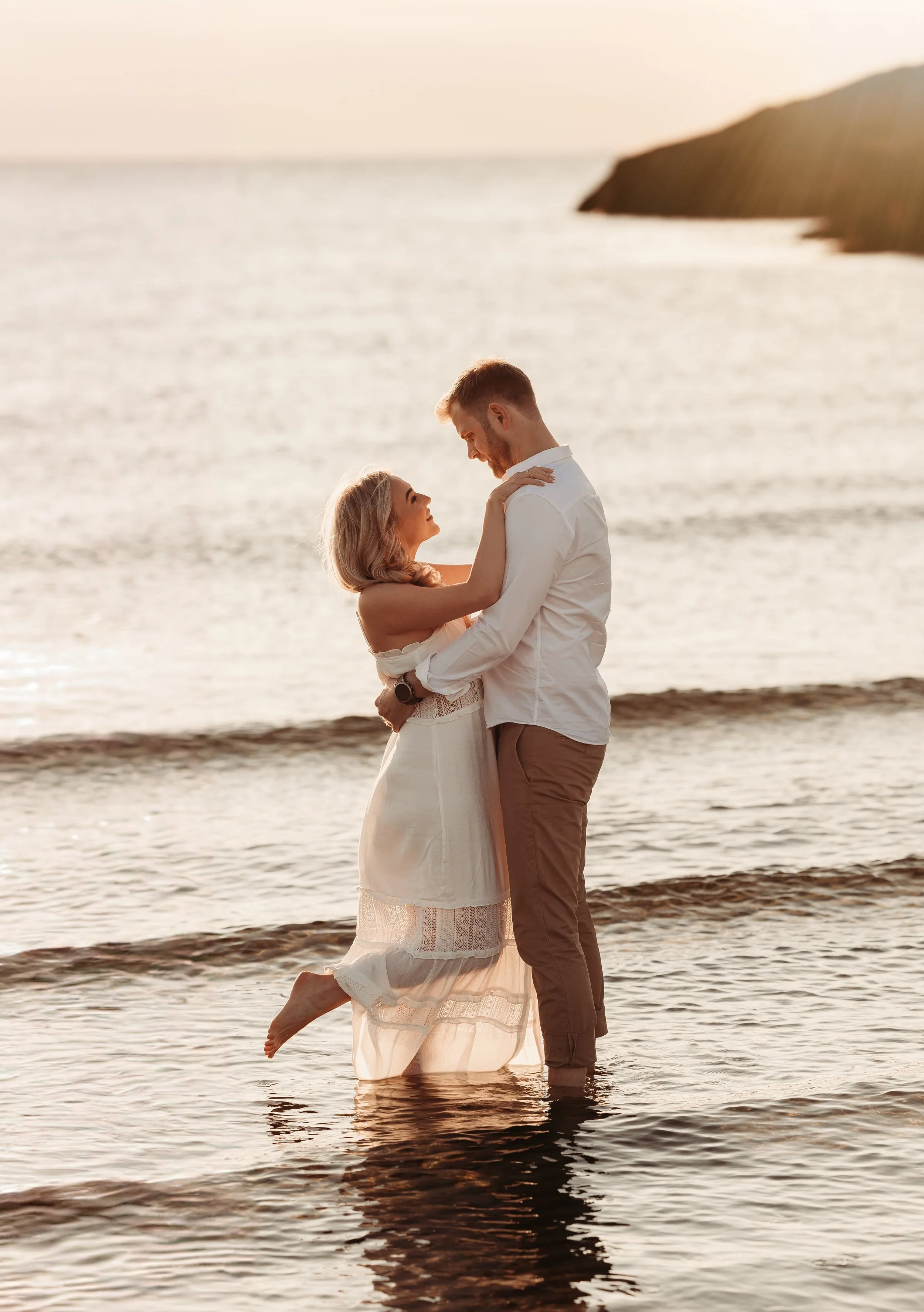 Et par står i vannkanten ved solnedgang, holder hverandre, med båtenes refleksjon i vannet.