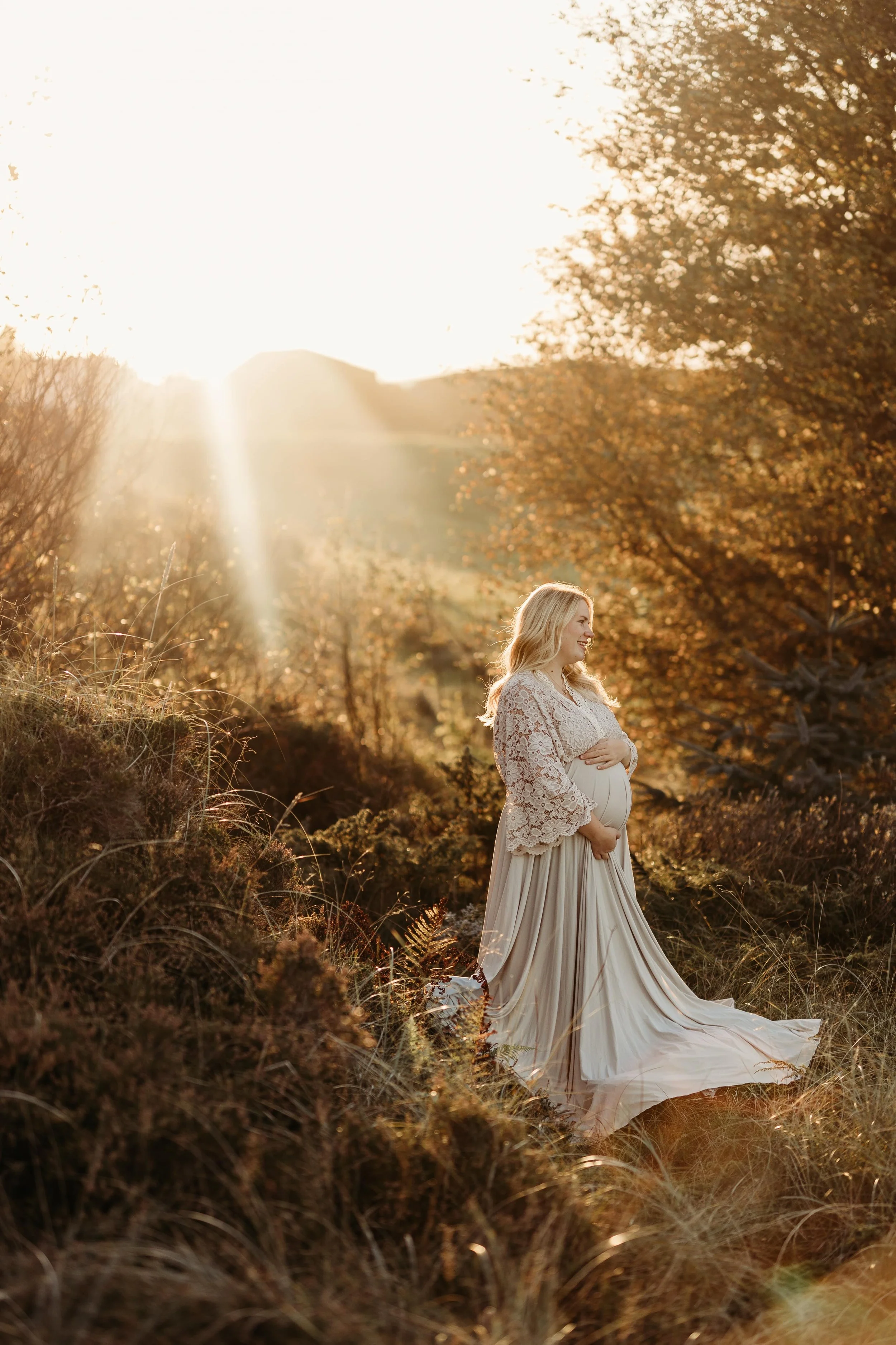 En gravid kvinne i en hvit kjole står i en landskap om høsten, omgitt av busker og trær med høstfargede blader, mens solen skinner inn fra venstre.