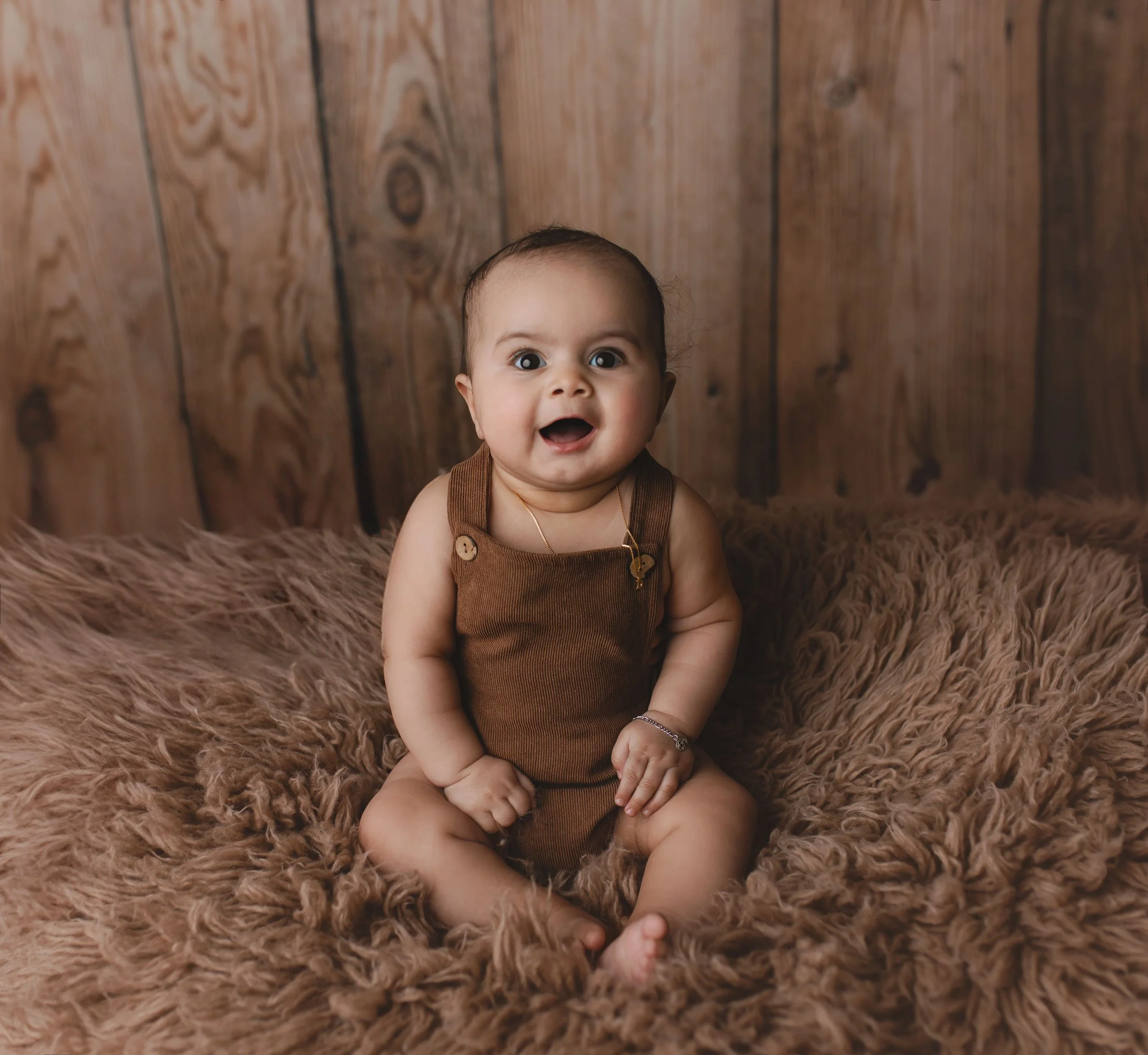 En baby som sitter på en myk, brun pels underlag med en trevegg bakgrunn. Babyen har på seg en brun overall og ser overrasket ut.