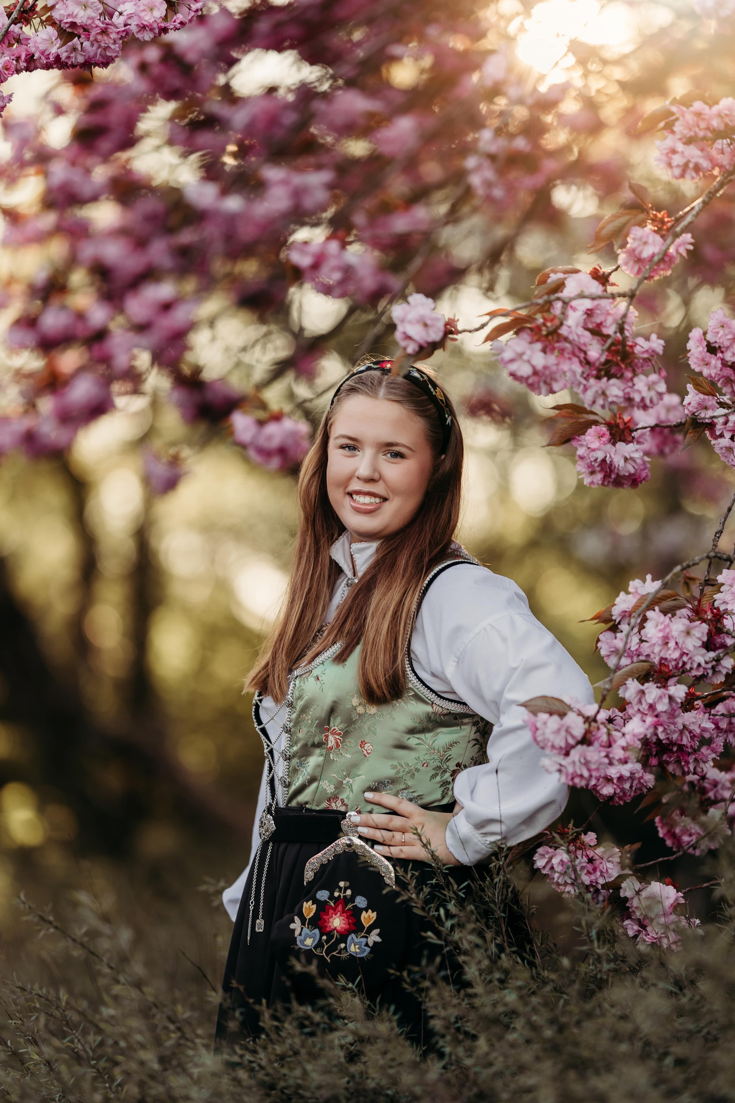 En smilende ung kvinne i tradisjonell norsk folkedrakt står blant blomstrende kirsebærtrær om våren, med sollys bak henne.