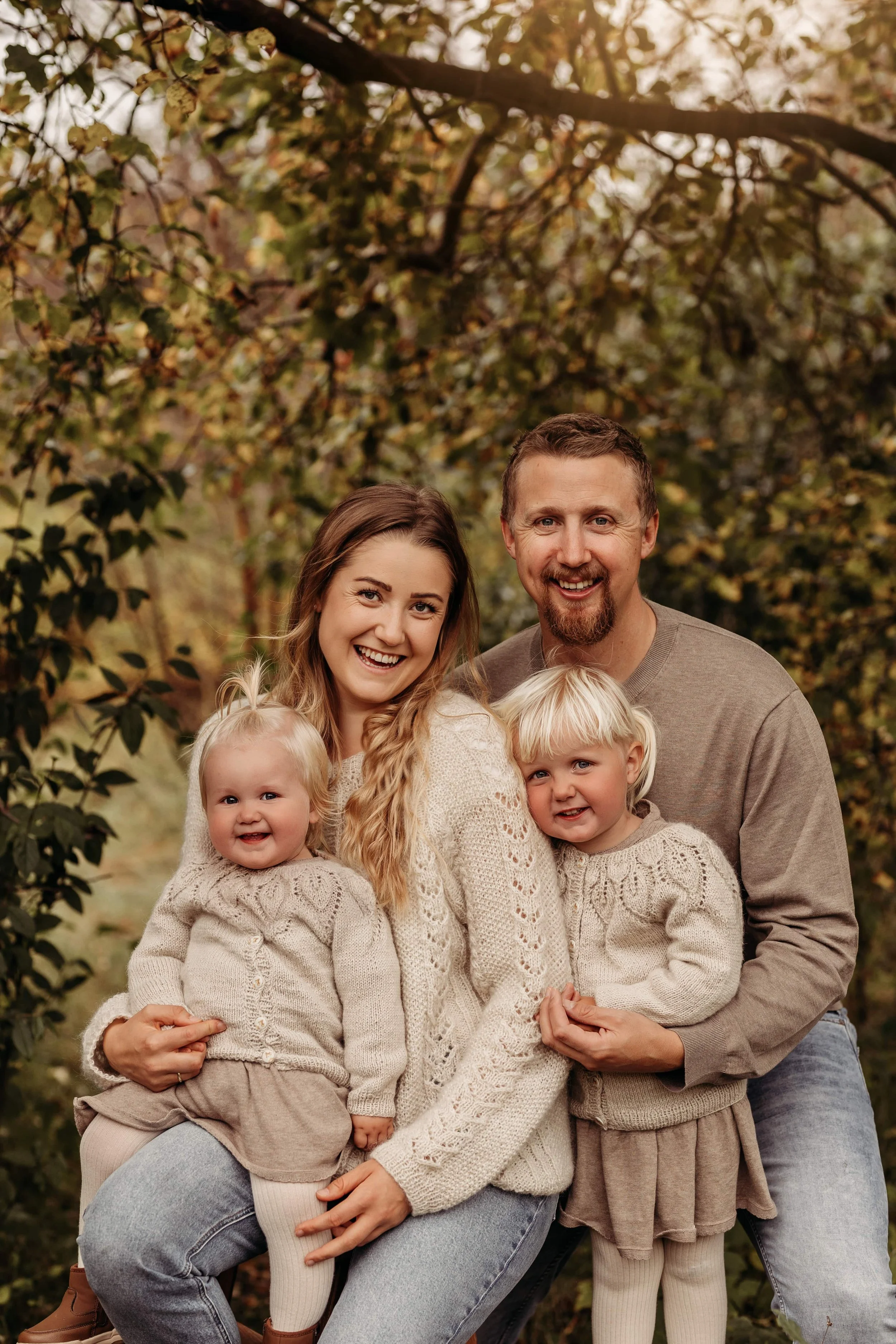 En familie med to voksne og to unge jenter smiler i en park med høstfarger.