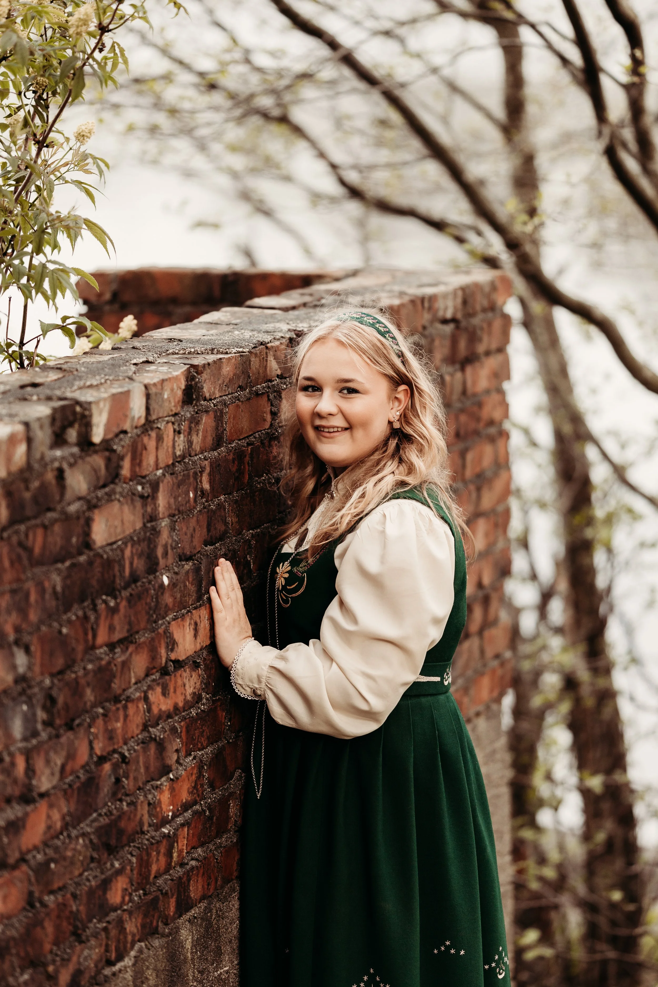 Ung jente i tradisjonell, grønn klesdrakt står med hånden på en gammel mur og smiler, omgitt av trær med lysegrønne blader.