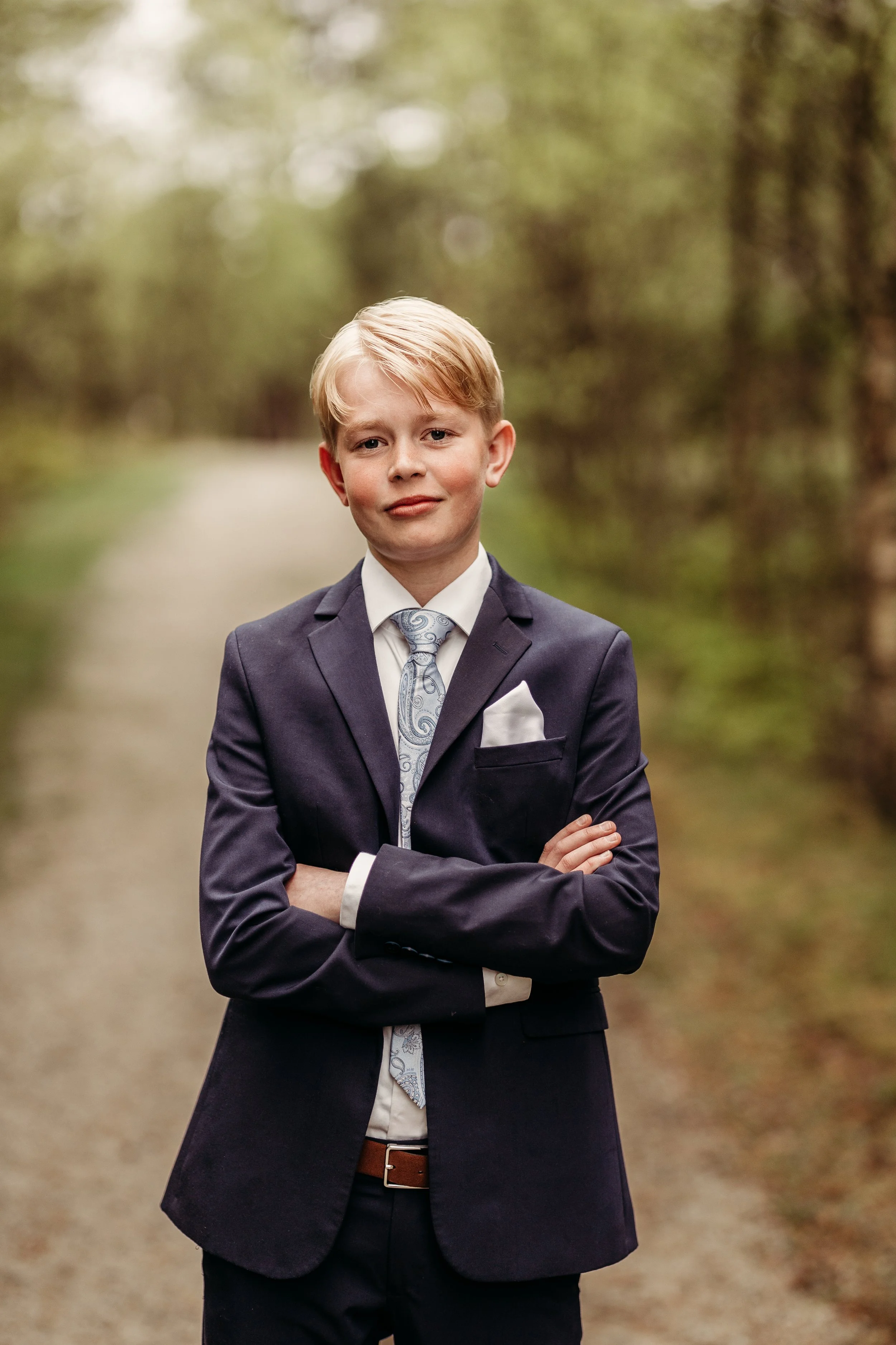 En ung gutt i dress står med armene crosset på en skogssti.