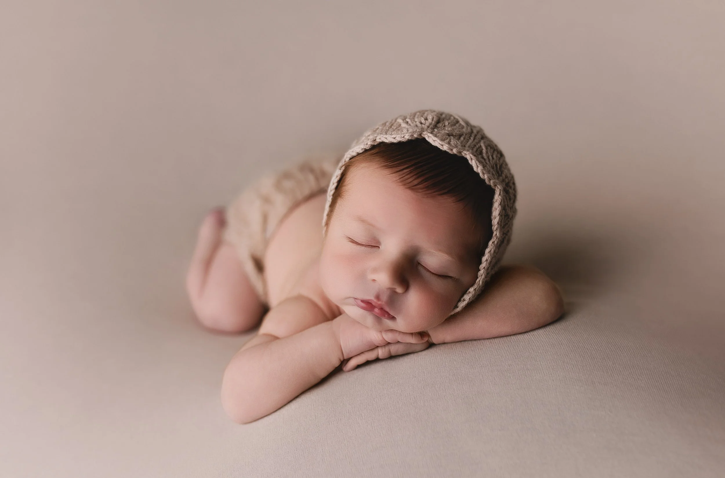 Søvnende baby med håret dekket av en strikket lue, hvilt på armen på en ensfarget beige bakgrunn.