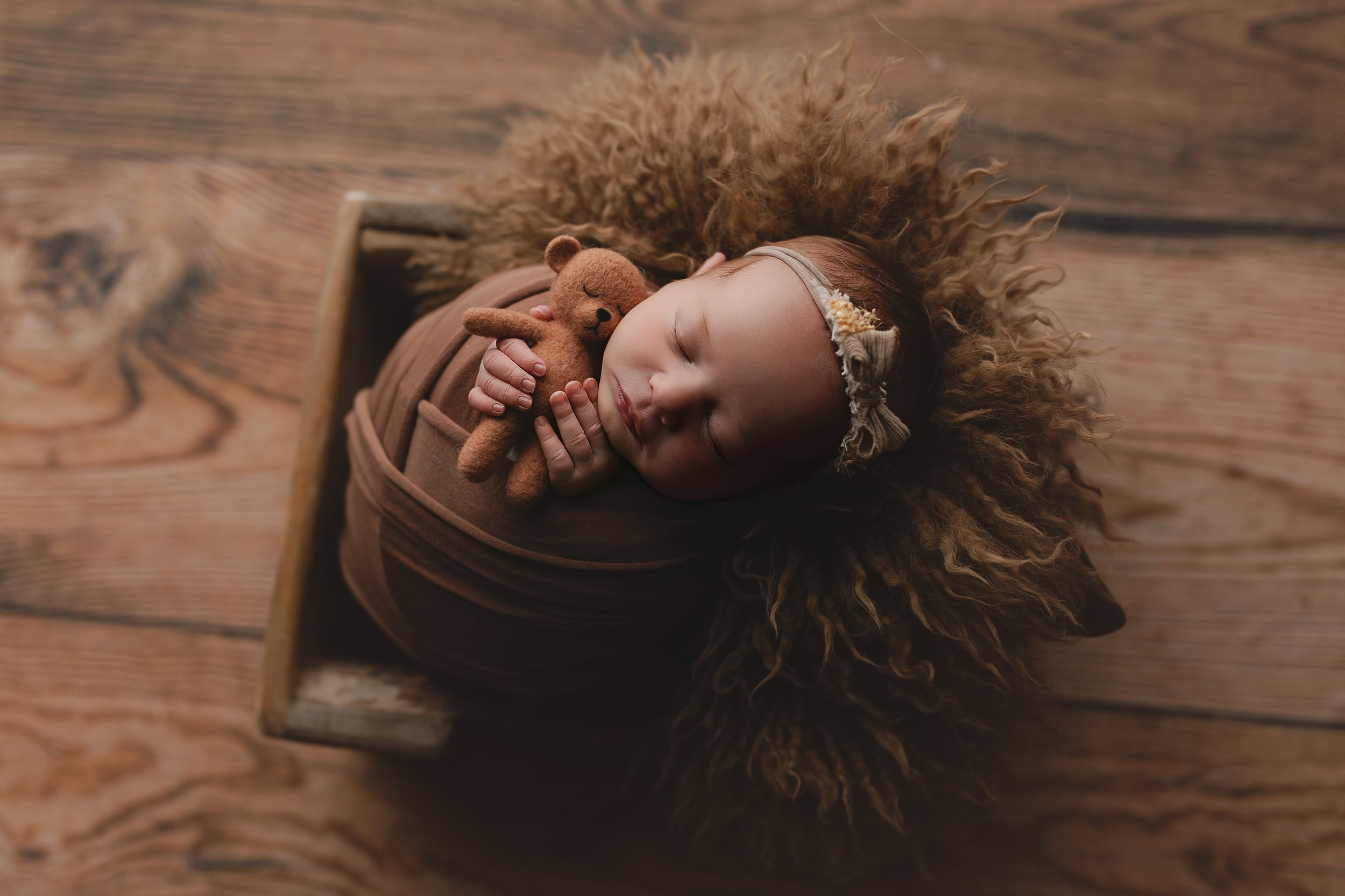 Søvnende baby med krøllete hår, inne i en treramme, bruker en beige hårbånd og holder en liten brun teddybjørn.
