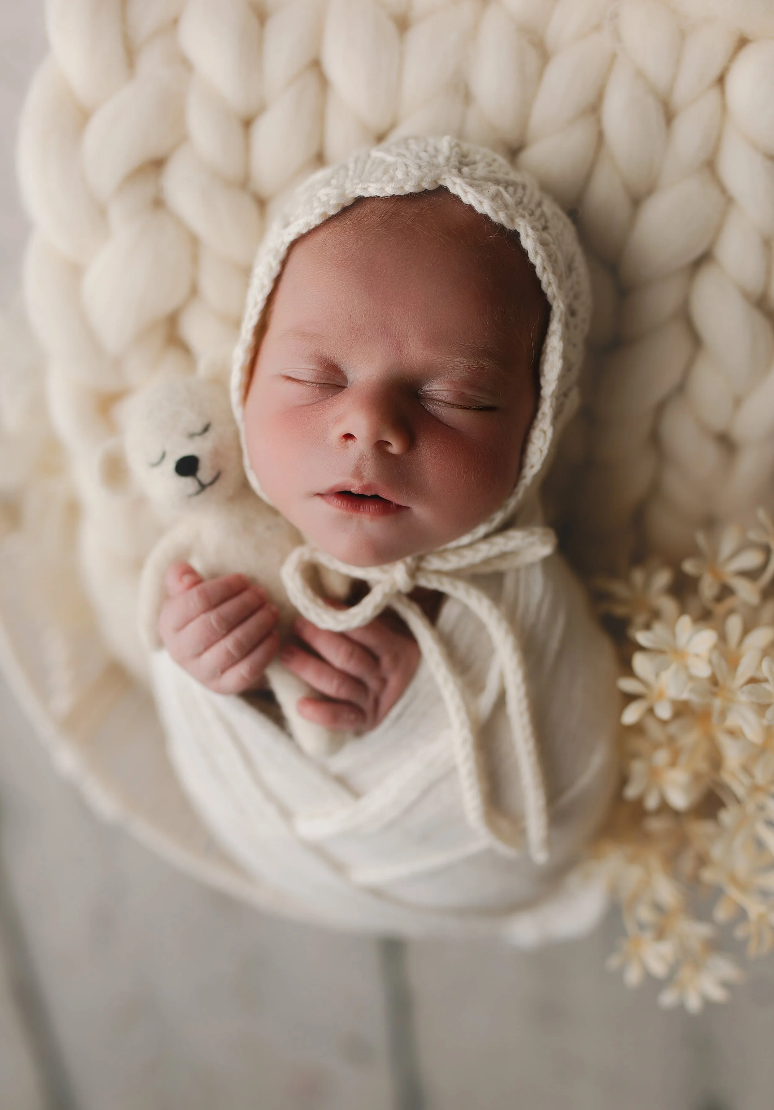 Småbarns baby med tett lue som sover, holder en liten bamse og ligger på en myk, kremfarget overflate med blomster rundt.