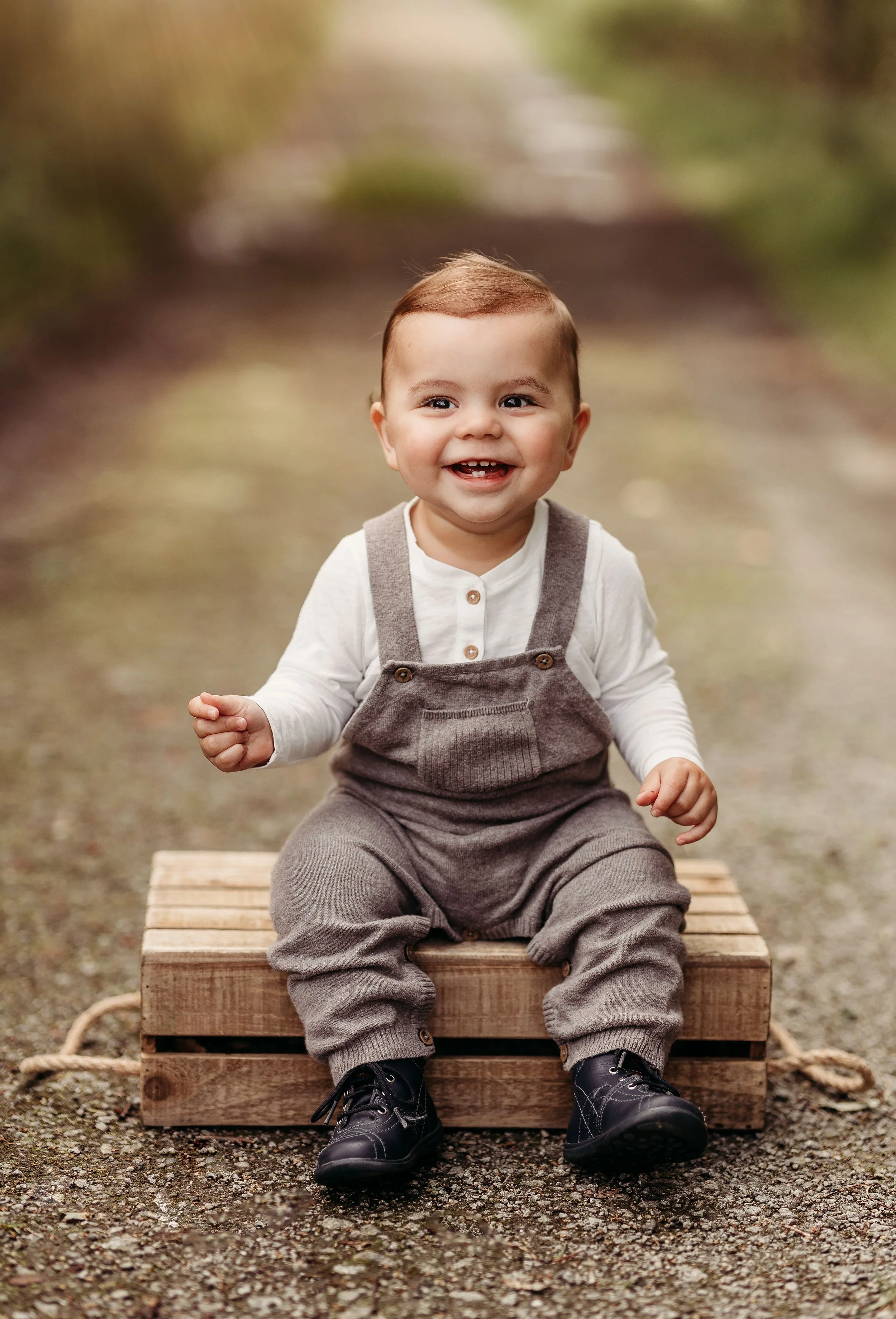 Smilende liten gutt som sitter på en trebenk på en grusvei i naturen, iført grå overalls og svarte sko.