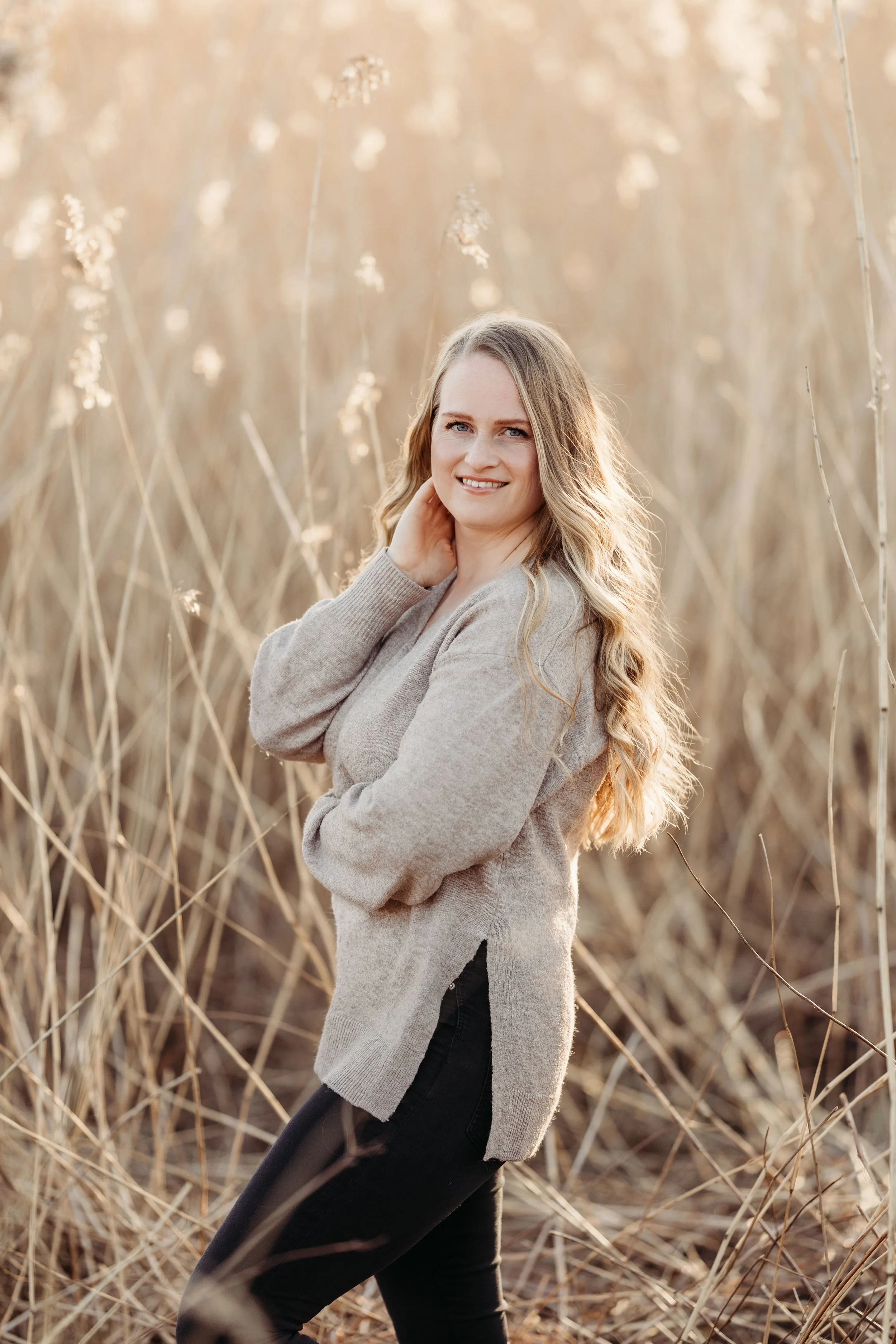 En kvinne med langt, bølget blondt hår smiler og poserer utendørs blant høye, tørre gresstrå om høsten.