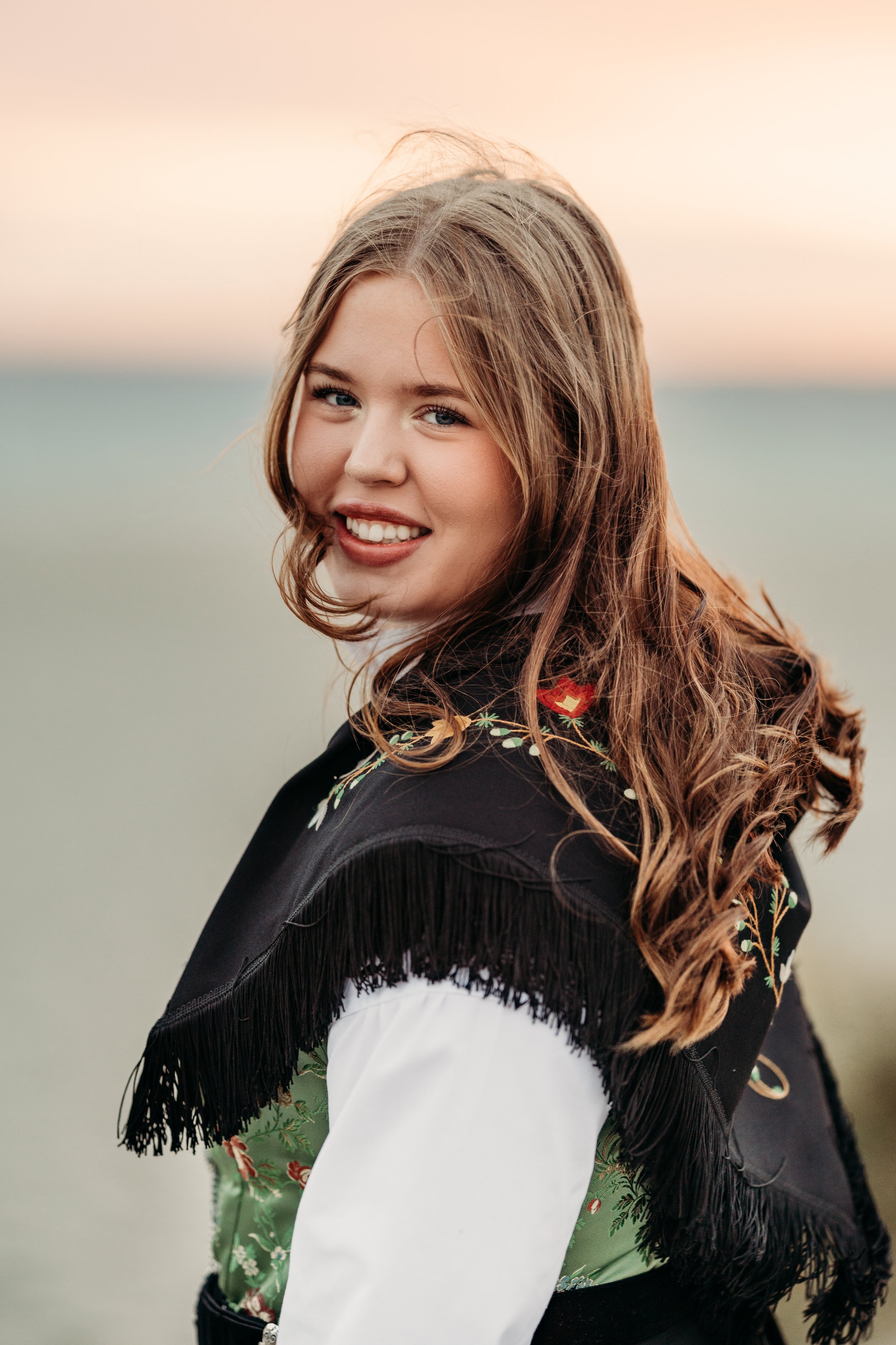 Et bilde av en smilende ung kvinne med langt, krøllete hår, som står utendørs ved solnedgang, iført en tradisjonell norsk bunad med blomsterbroderi og frynser.