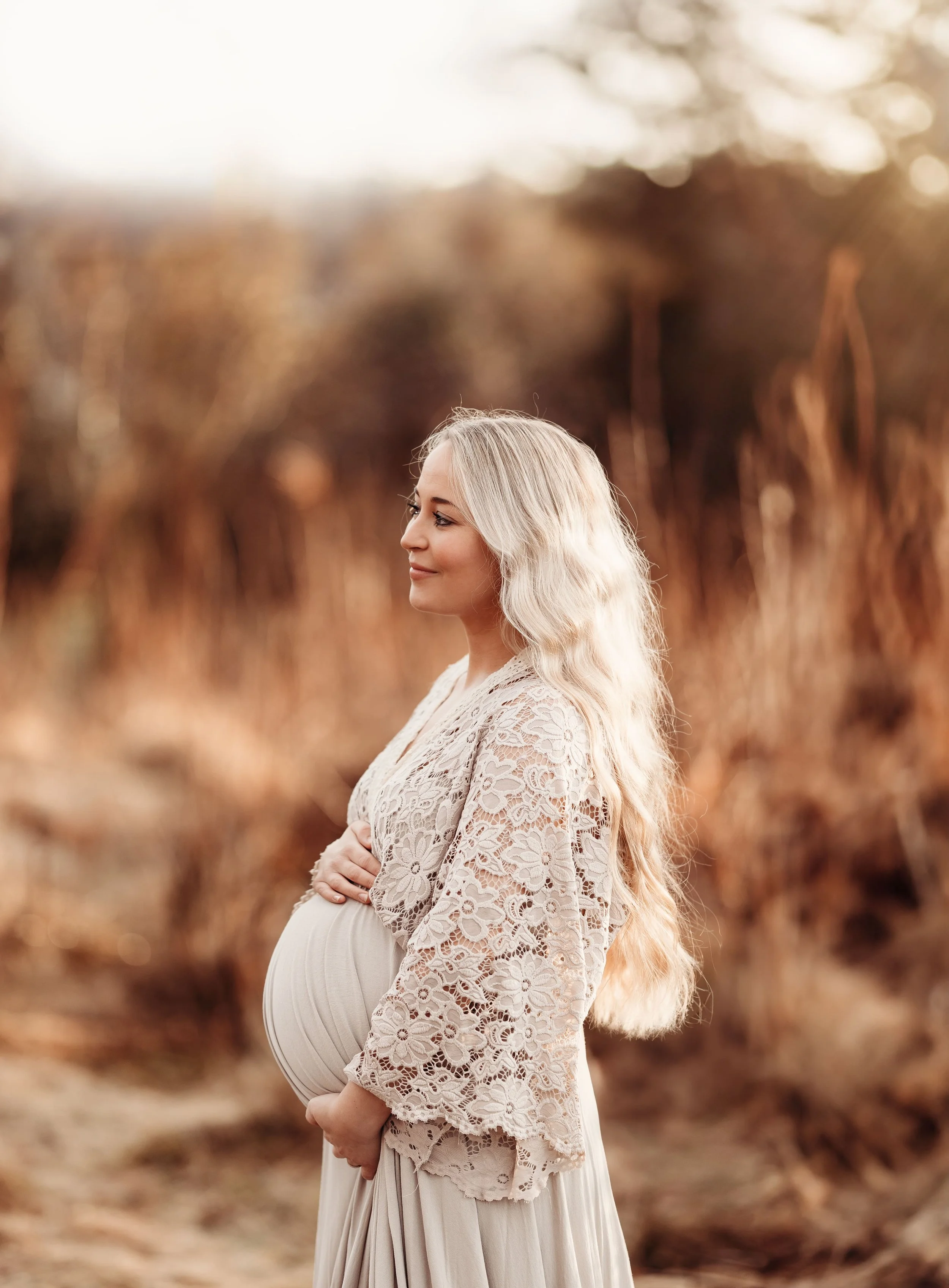 En gravid kvinne ikledd en kremfarget kjole og blondt, langt, bølget hår står i en åker med gult gress, og solen skinner i bakgrunnen.