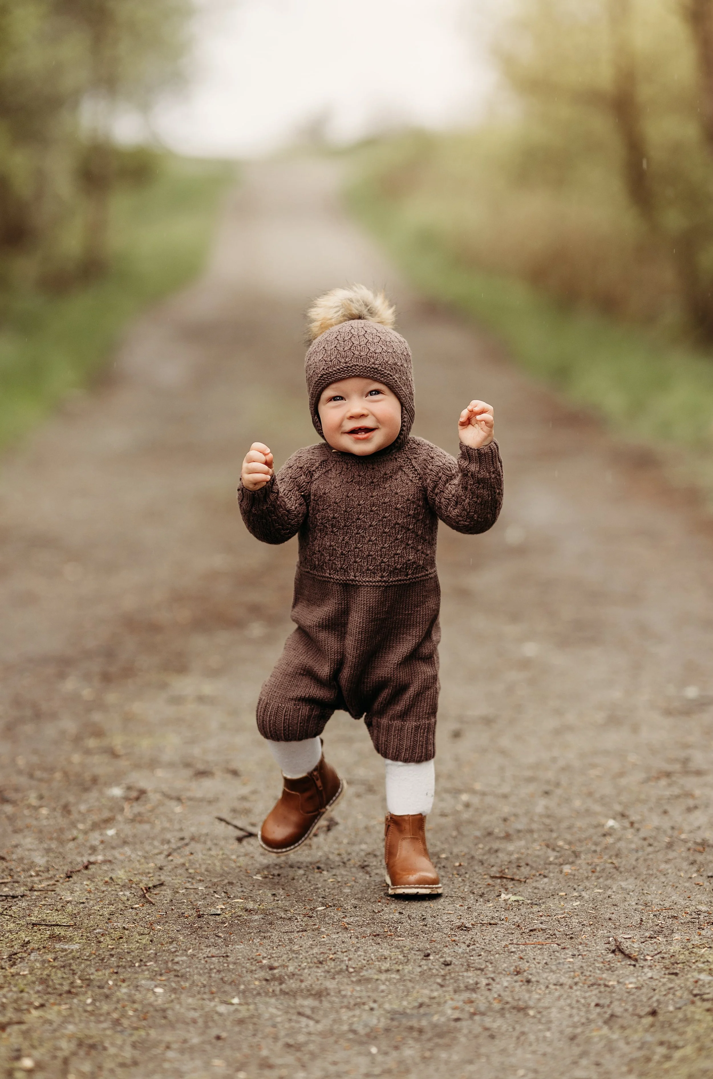 Liten gutt som løper på en skogssti med smil og varme klær, inkludert brun jakke, hat og votter.