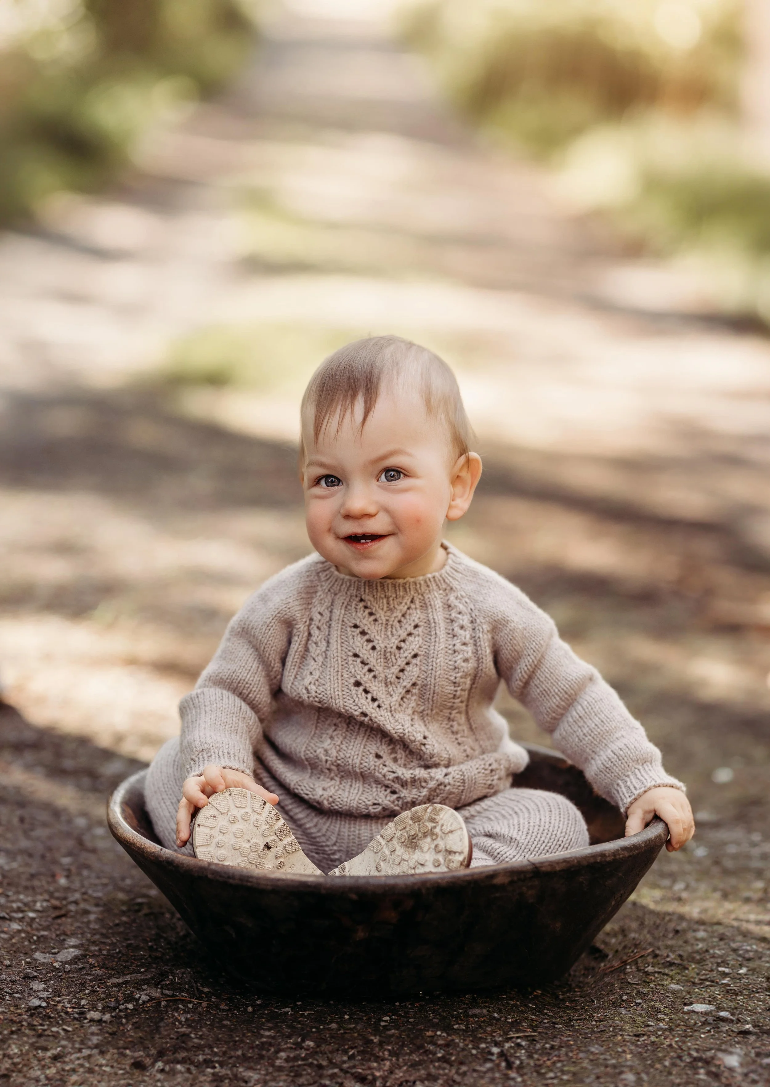 Liten gutt sitter i en svart bøtte utendørs, iført en beige genser, med solskinn og skog i bakgrunnen.