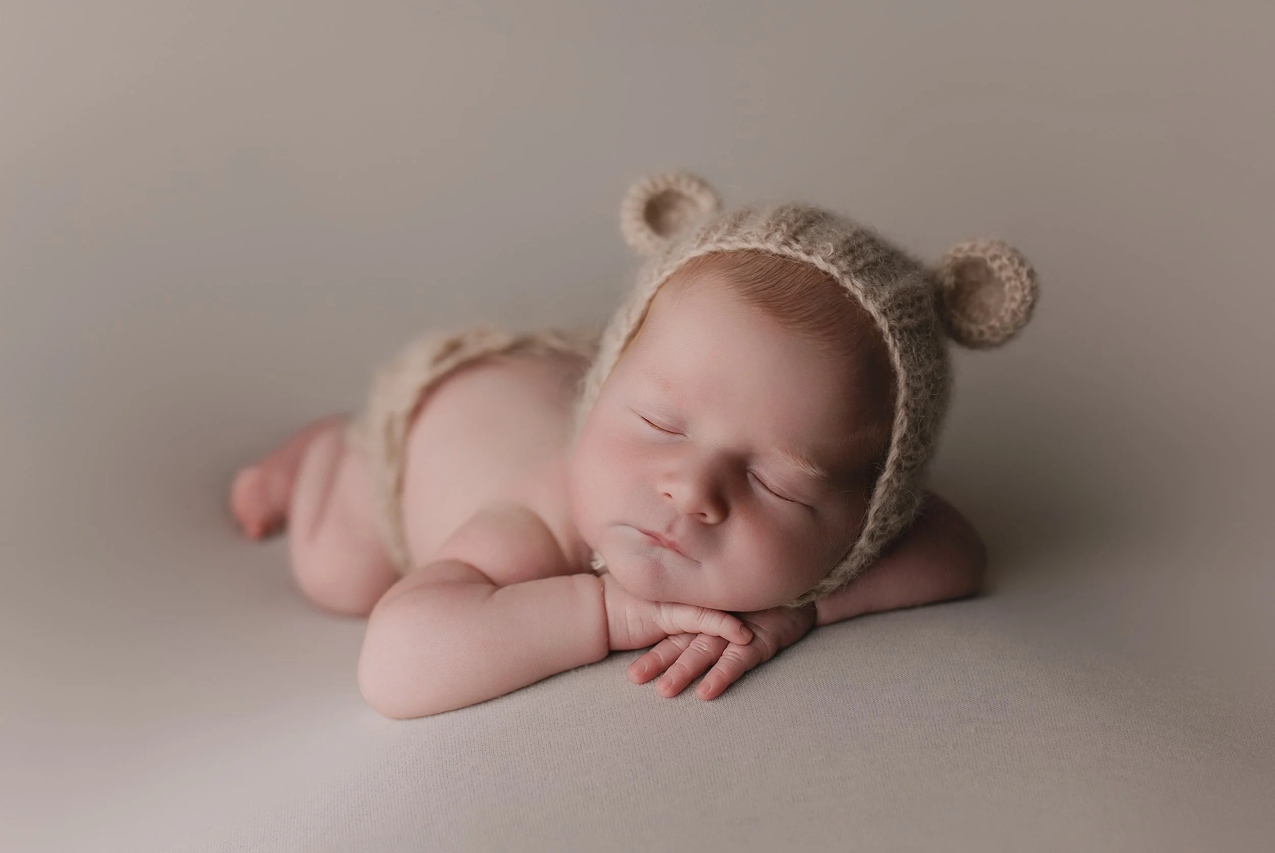 Søvnende baby i beige hue med ørene og topp i form av bjørneører, liggende på mage på en myk overflate.