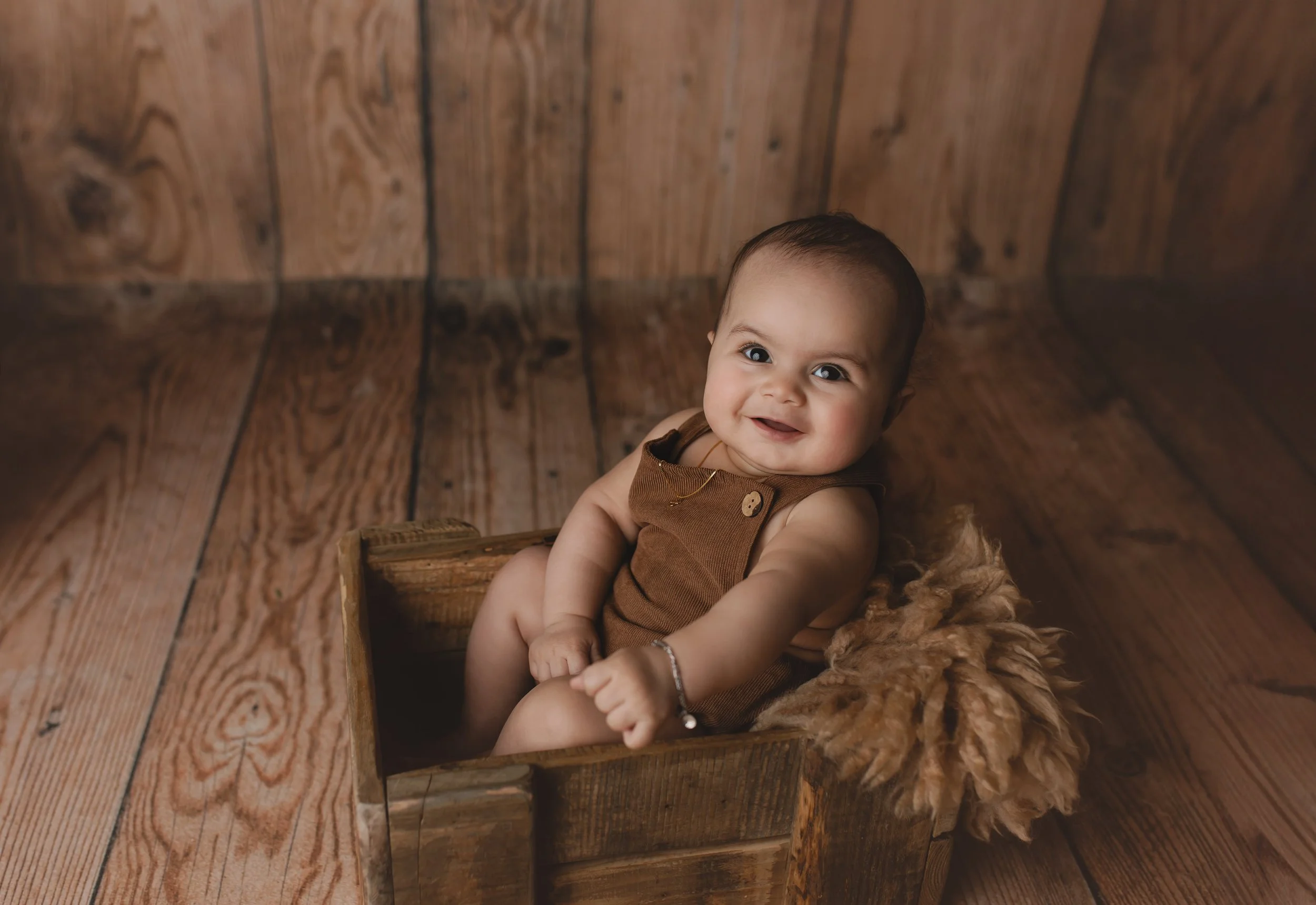En søt baby i brunt antrekk sitter i en liten trekasse på en trebjelkelag. Babyen smiler og ser mot kamera, omgitt av en fluffy brungardin.