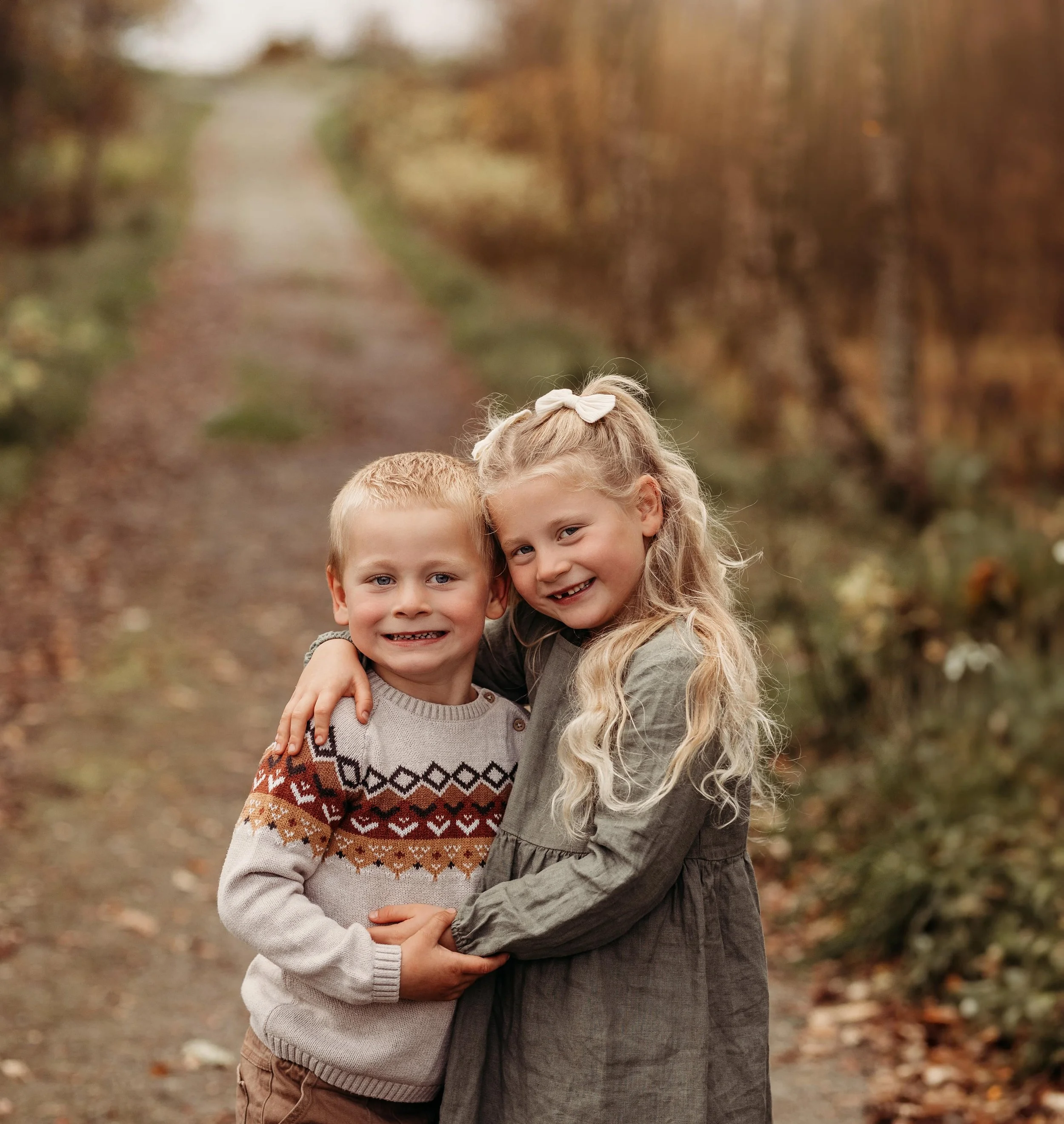To unge barn, en gutt og en jente, står tett sammen på en skogssti, omgitt av høstfargede trær. De smiler og omfavner hverandre.