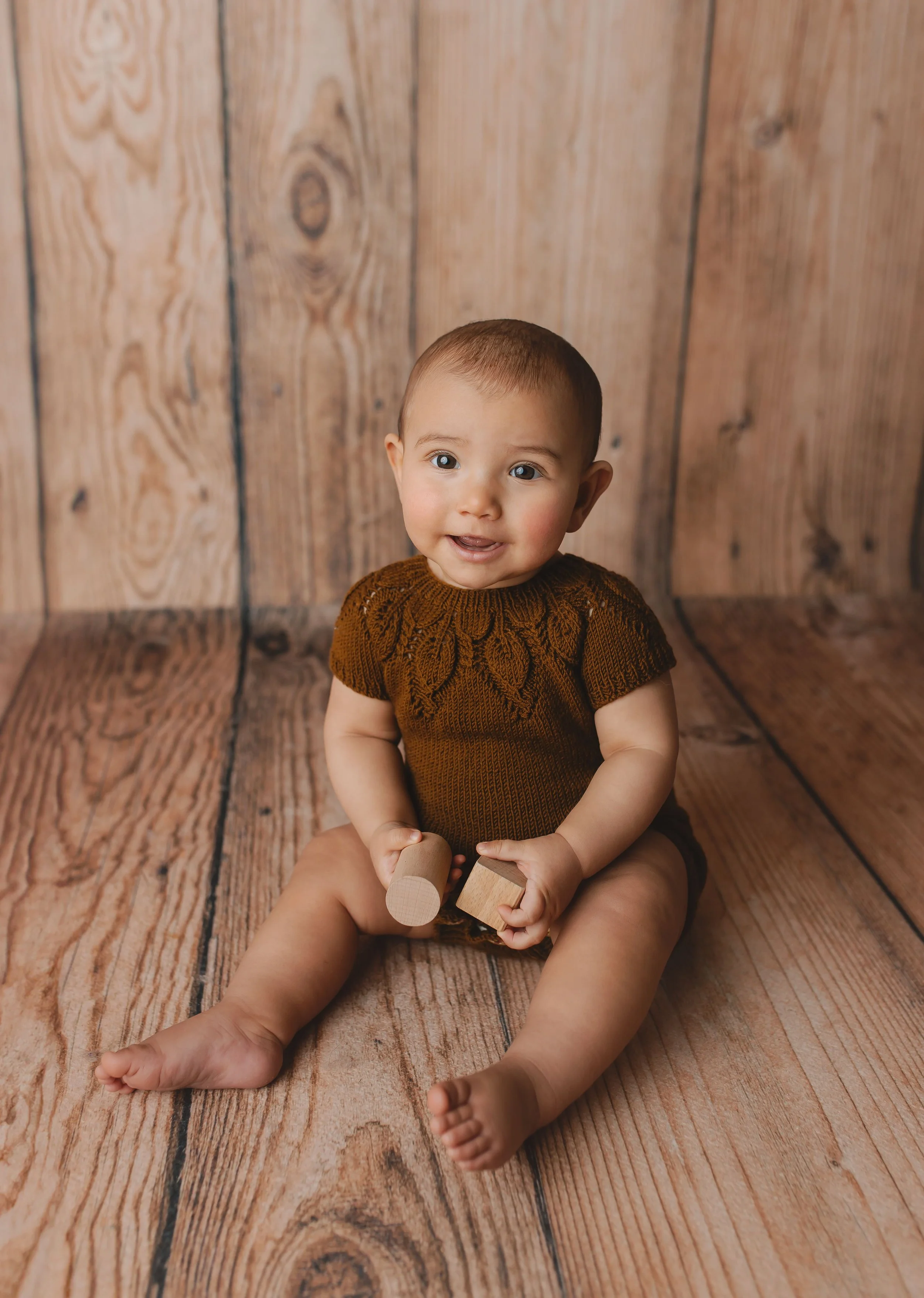 En baby sitter på en trebord bakgrunn, iført en brunt strikkegenser og holder to treklosser, smiler mot kamera.