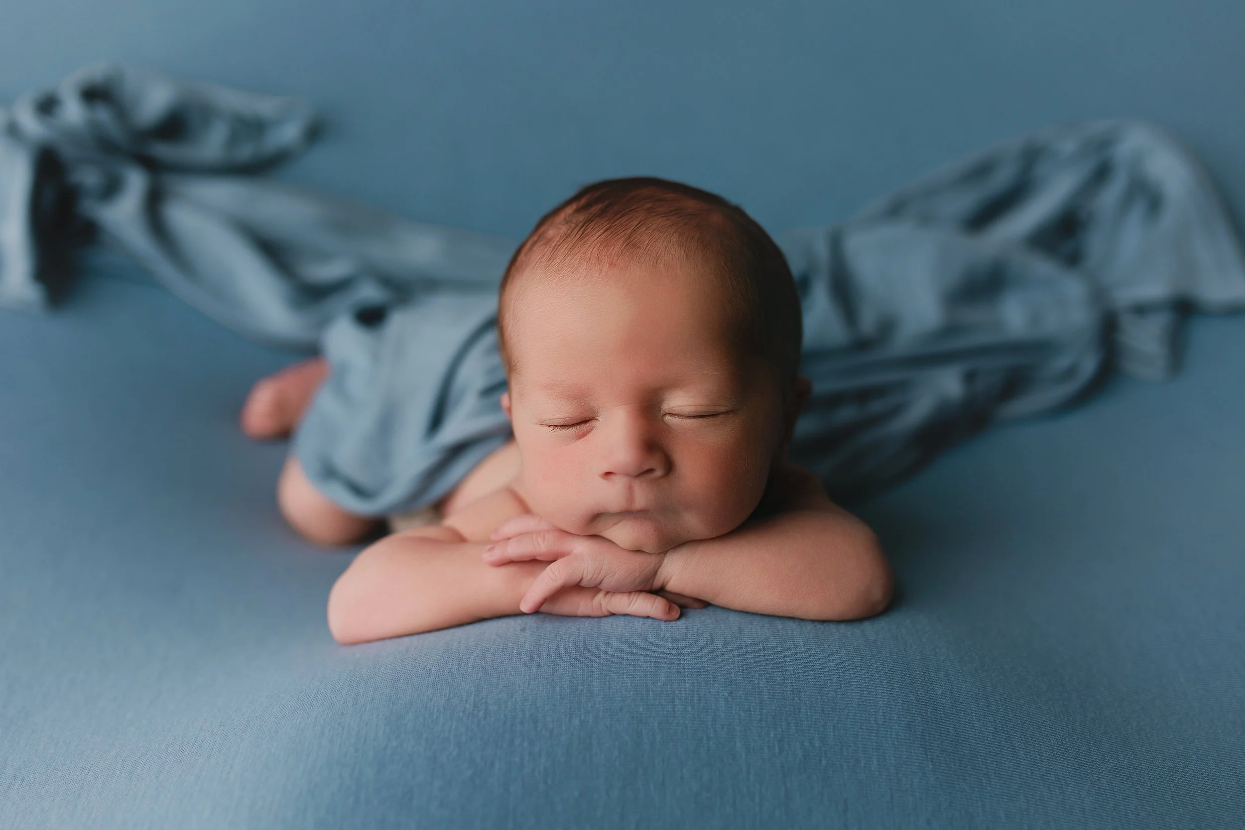 Nydelig nyfødt baby som sover med hodet hvilende på armene på en blå overflate, kledd i blått.