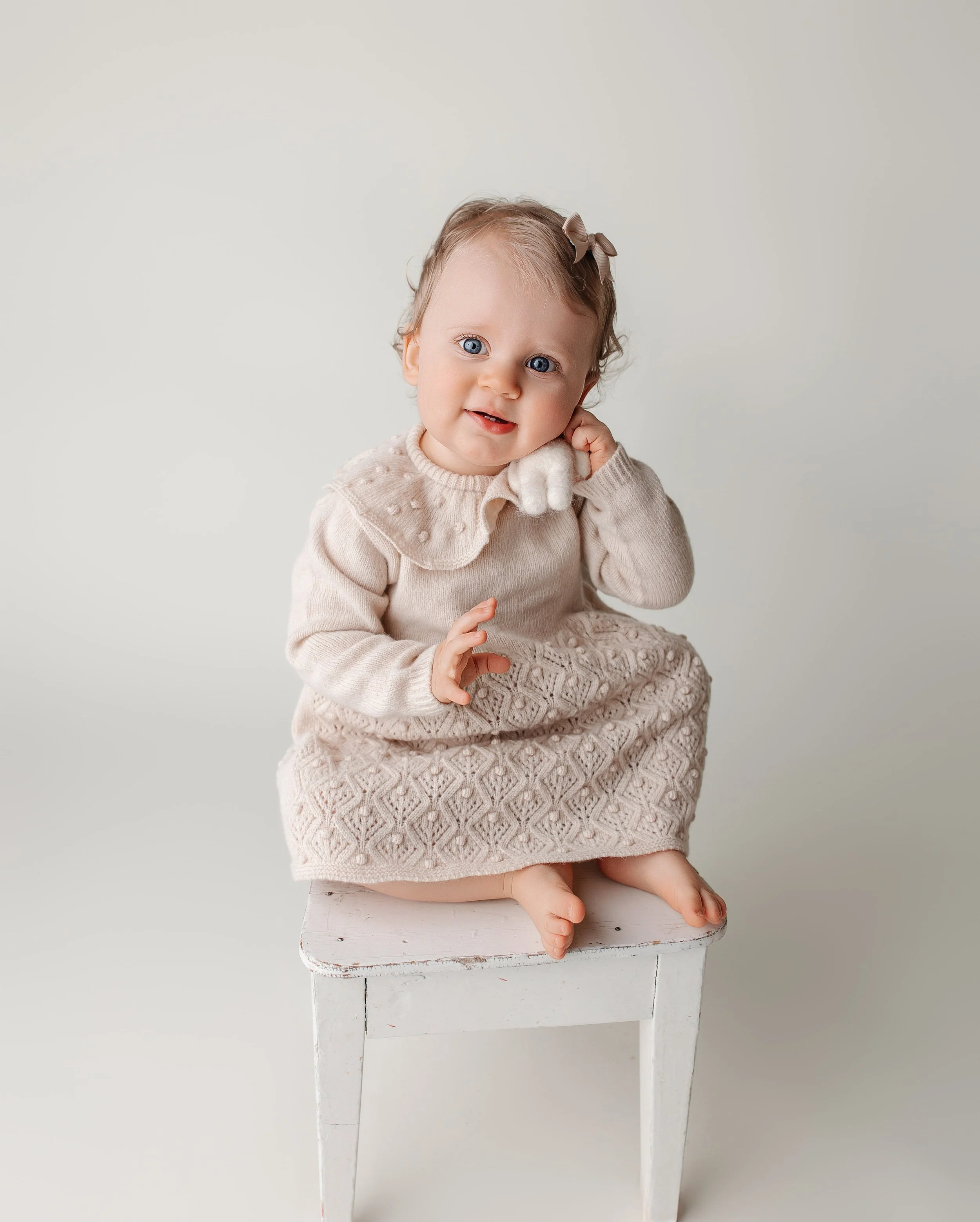Et søtt lite barn med blå øyne, iført lys beige genser med pelskrage, og en matchende lang kjole, sitter på en hvit trestol mot en enkel lys bakgrunn.