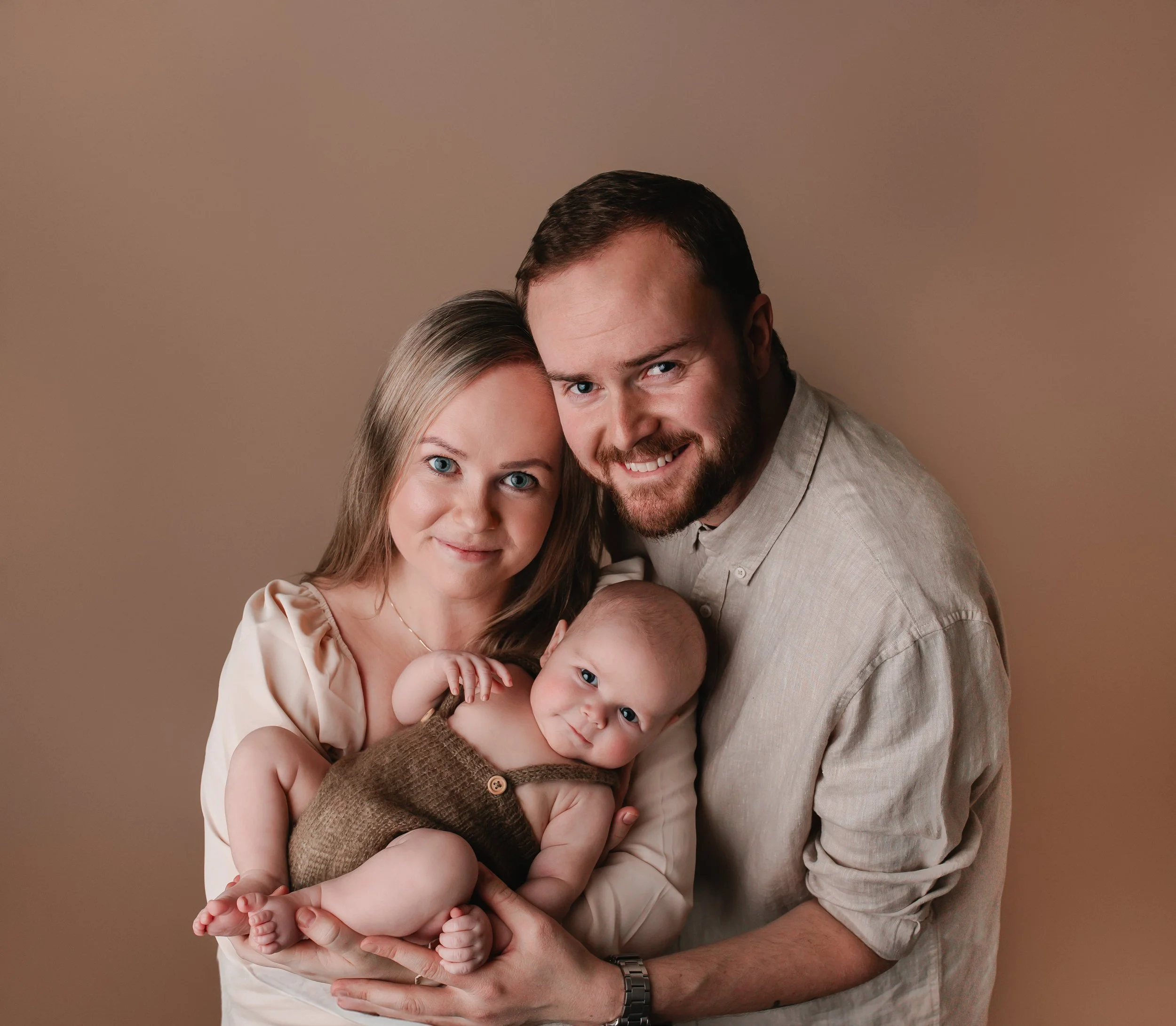 Et smilende familiebilde av en kvinne, en mann og en baby mot en beige bakgrunn.