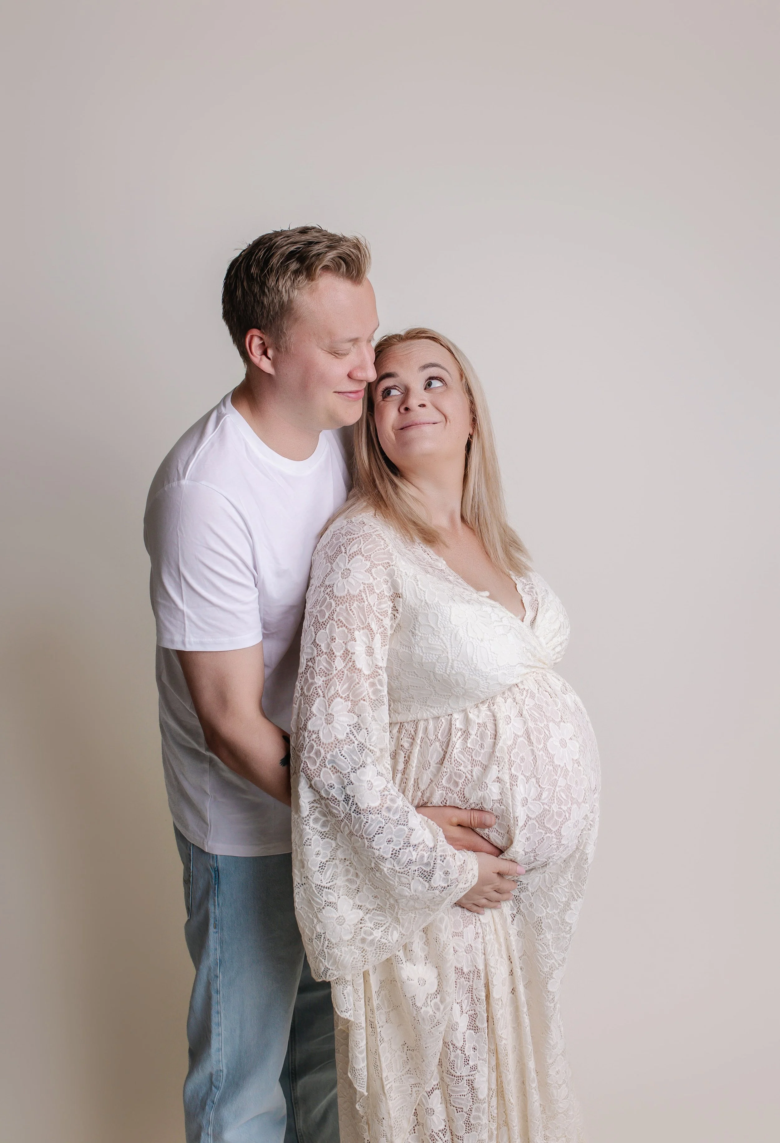 En gravid kvinne i kremfarget blonder kjole, står foran en mann i hvitt T-skjorte, med et smilende ansiktsuttrykk.