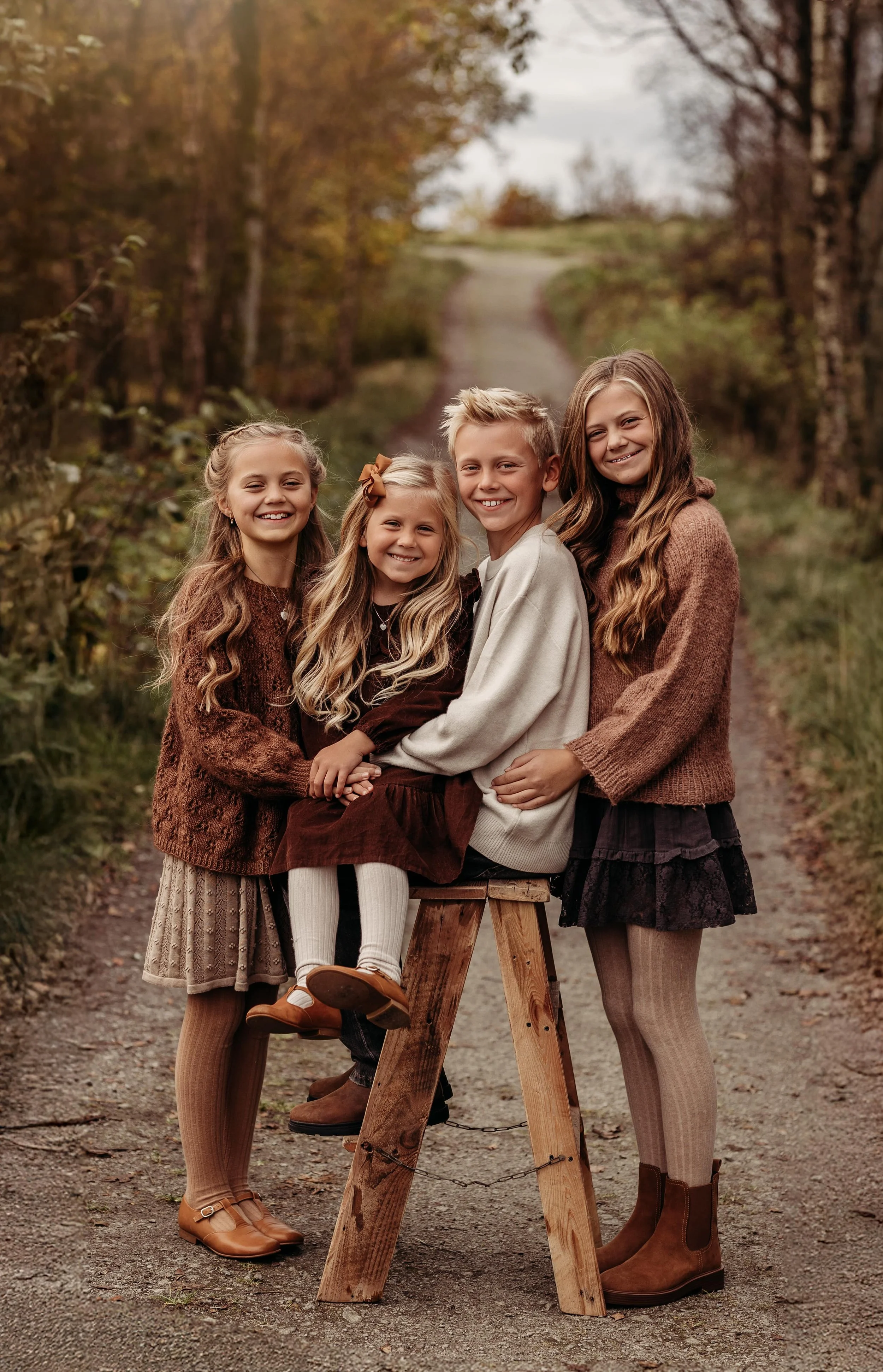 Fire barn og en ungdom står sammen utenfor i en skogsti, smilende og kledd i høsttoner.