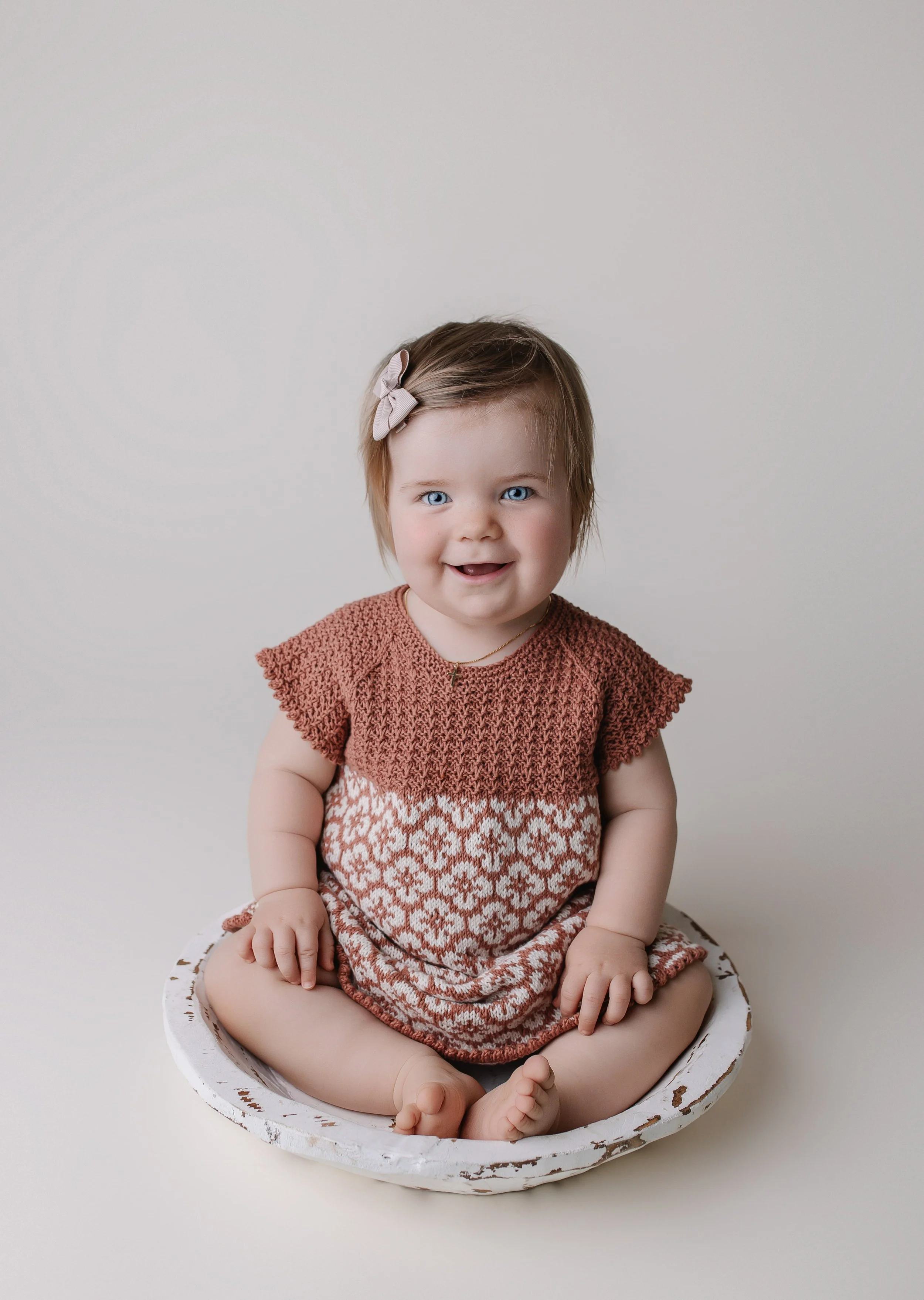 En baby med blå øyne, skulderlangt brunt hår med en rosa hårspenne, sitter krysslagt på en hvit, malt trefat mot en lys bakgrunn, iført en rustorange og hvit stripete kjole.