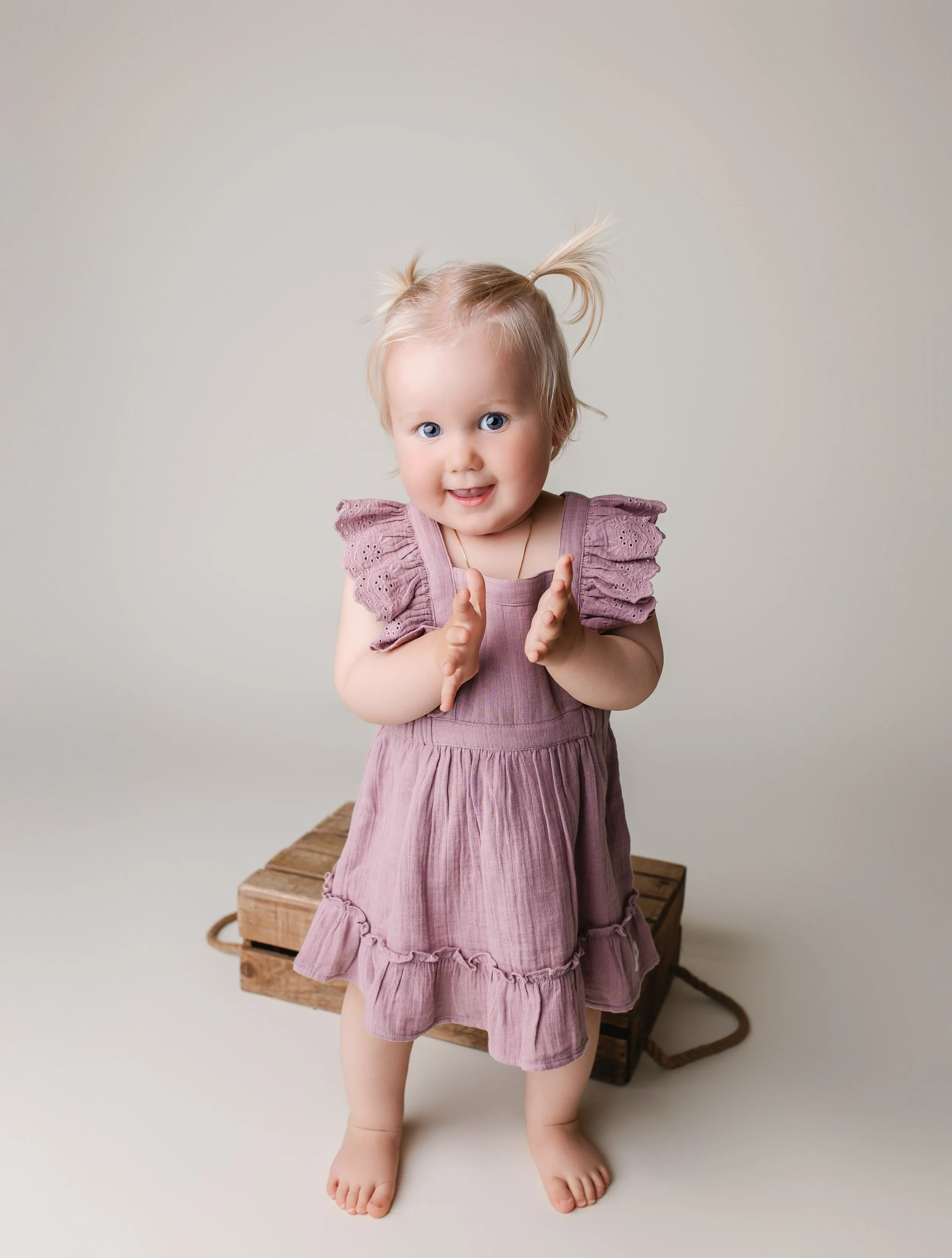 En liten jente med blondt hår og blå øyne står foran en enkel, lys bakgrunn. Hun har på seg en lilla kjole og er uten sko, med et smilende ansikt og hendene løftet frem som om hun skal klappe.