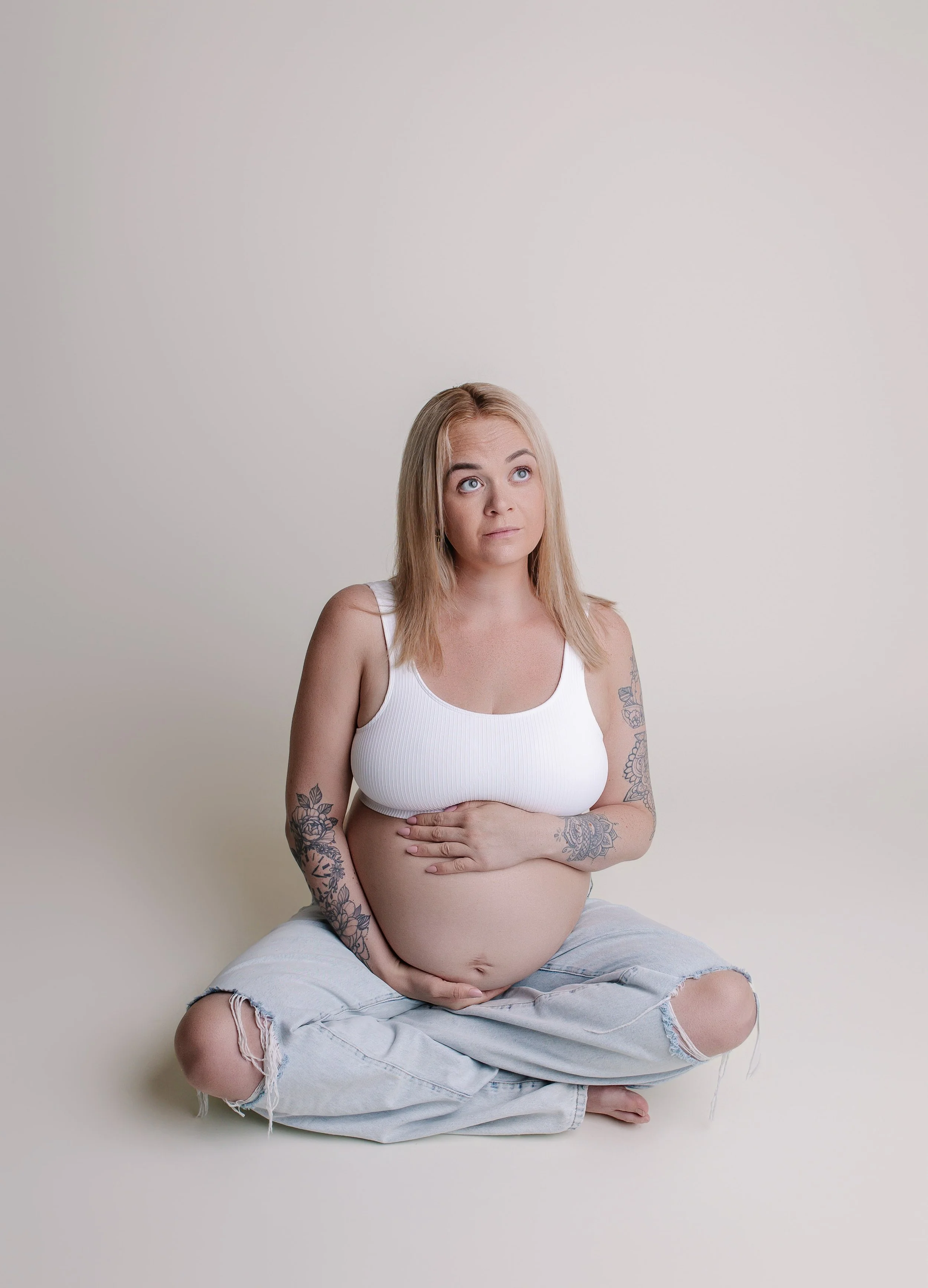 Gravid kvinne med tatoveringer sitter korslagt mot en lys bakgrunn, iført hvit top og slitte jeans.