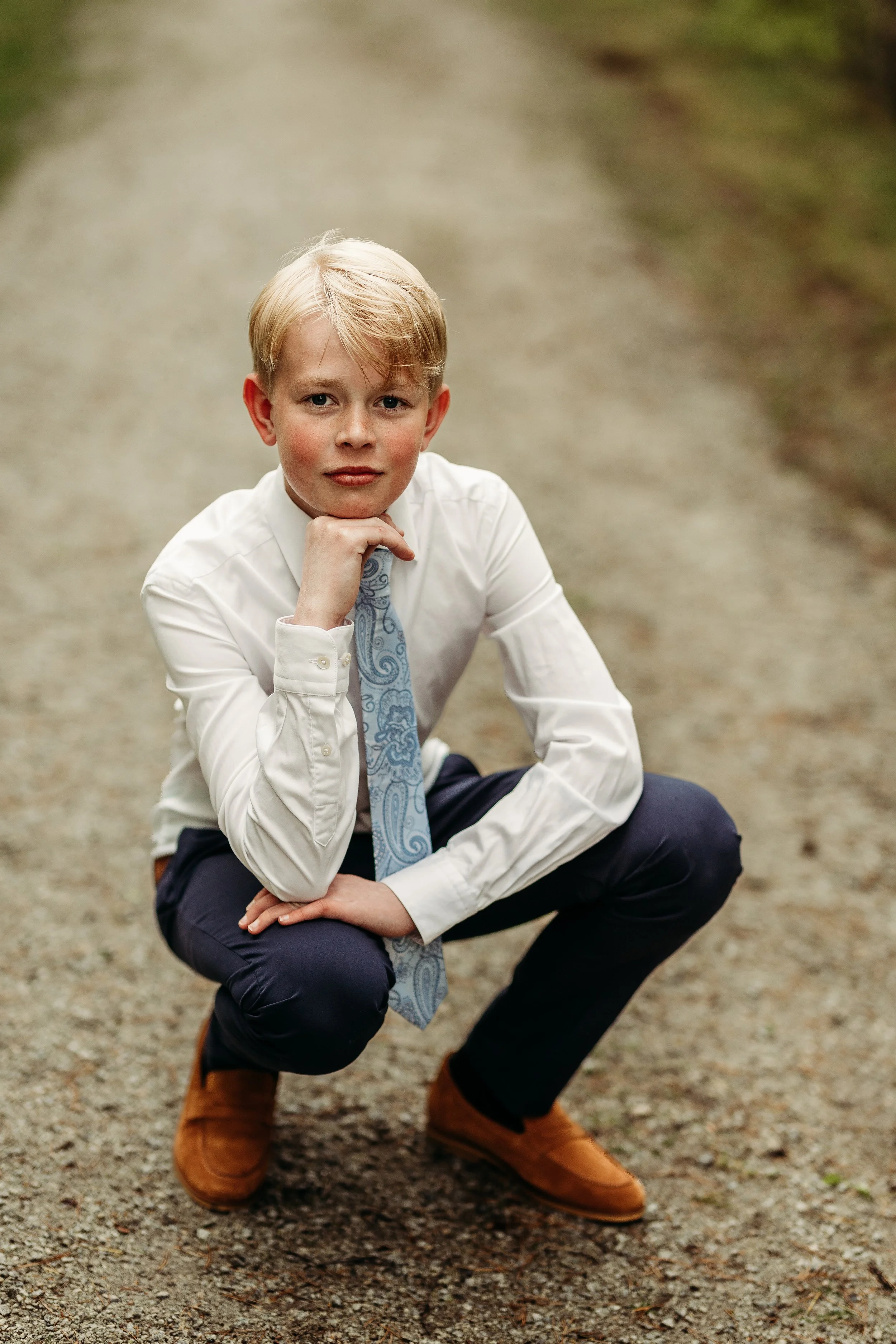 En ung mann med blond hår i dress, knepper nede på en grusvei i en utendørs setting.