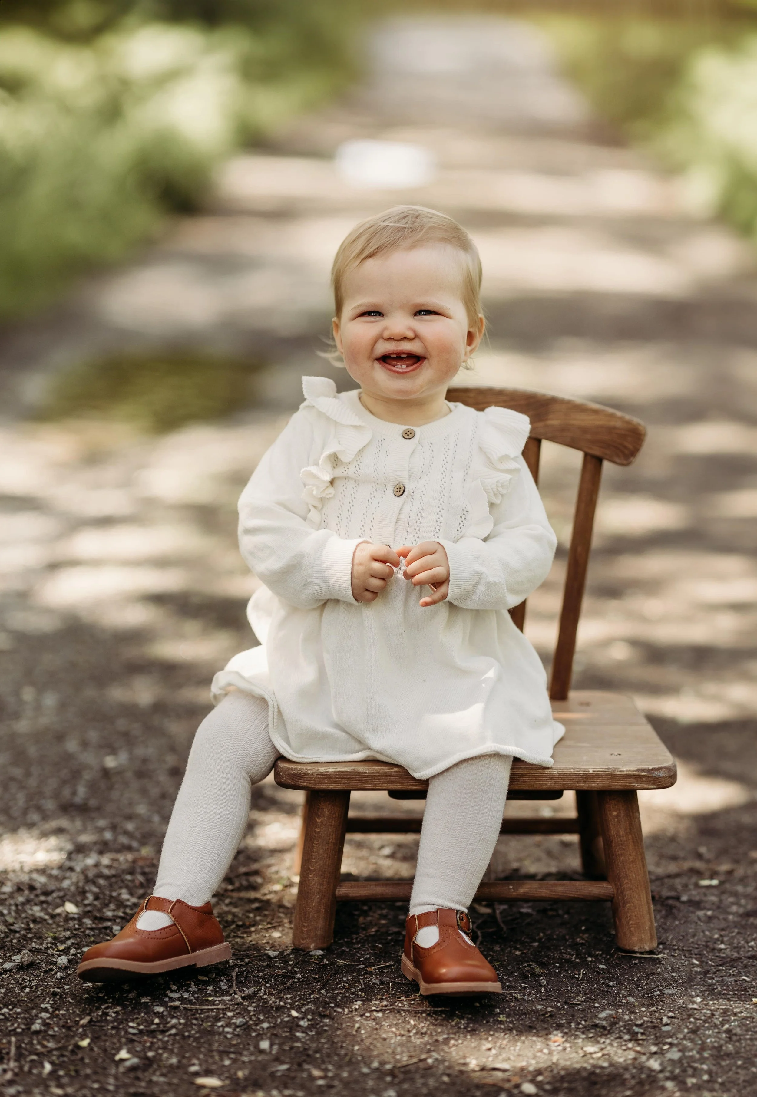 Liten jente med lett hår sitter på en trestol utendørs i en skogssone iført en hvit kjole og brune sko, smilende.