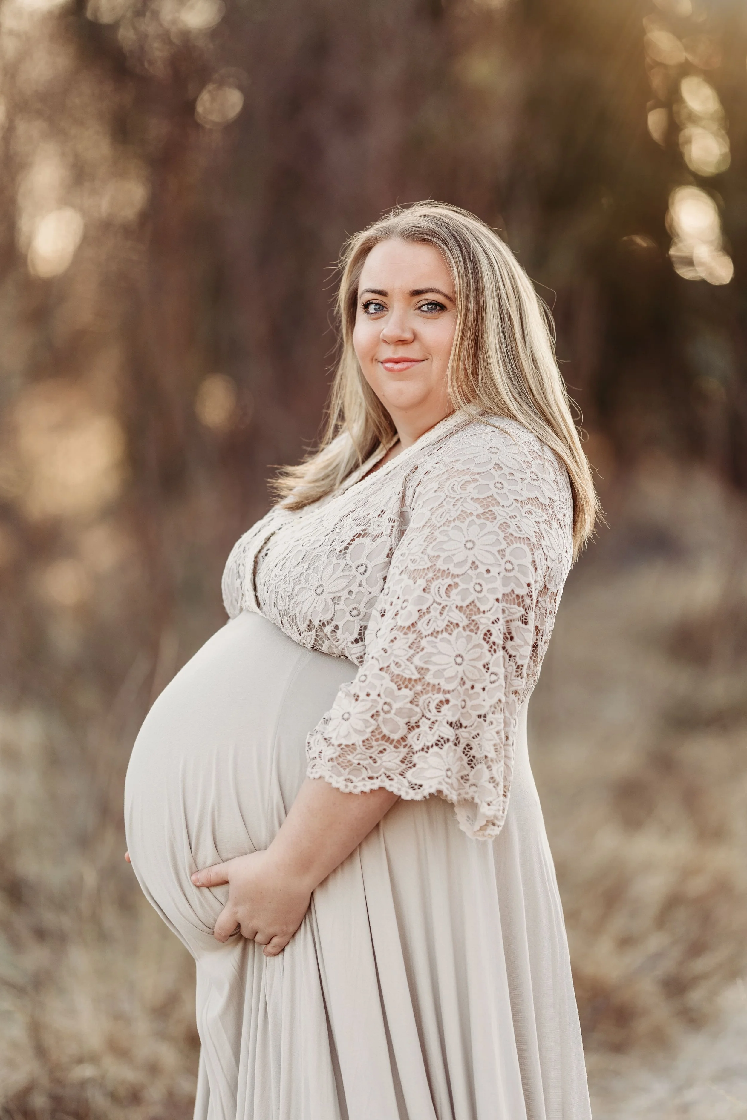 Gravid kvinne i lang lys kjole og blonderjakke står i utendørs landskap, ser mot kamera, med bløtt fokus bakgrunn.