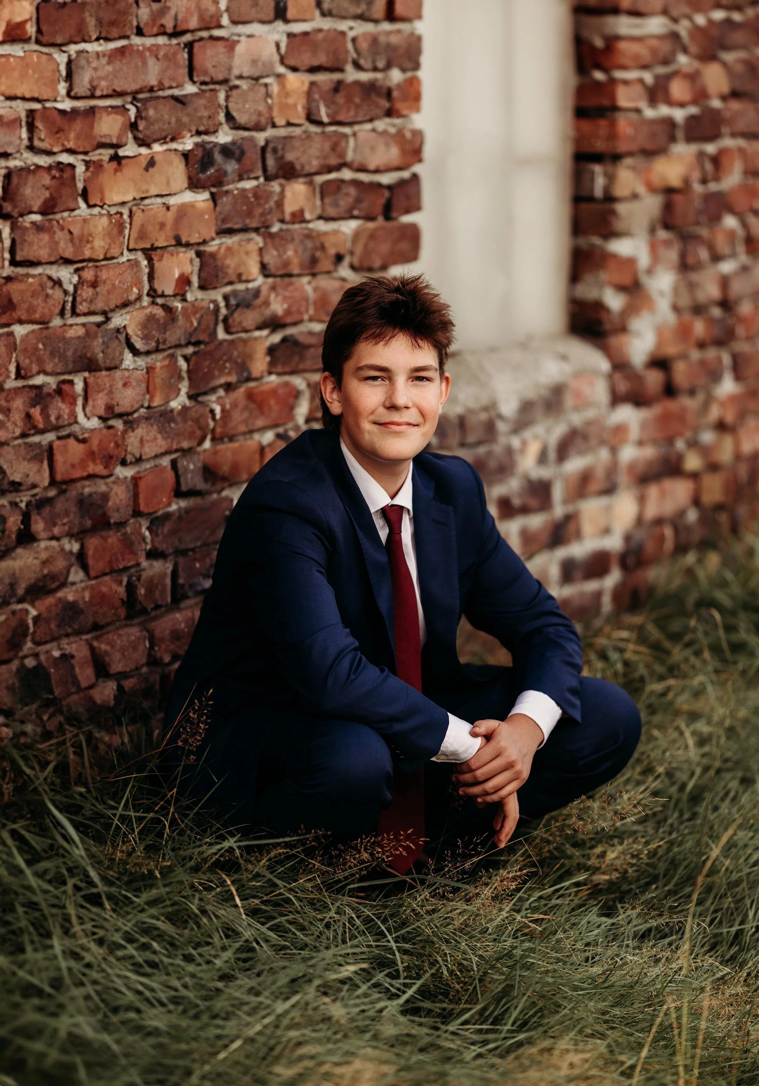 Ung mann i formell dress, sitte på knær i gress ved en mur av murstein, med en oppreist holdning og et vennlig smil.