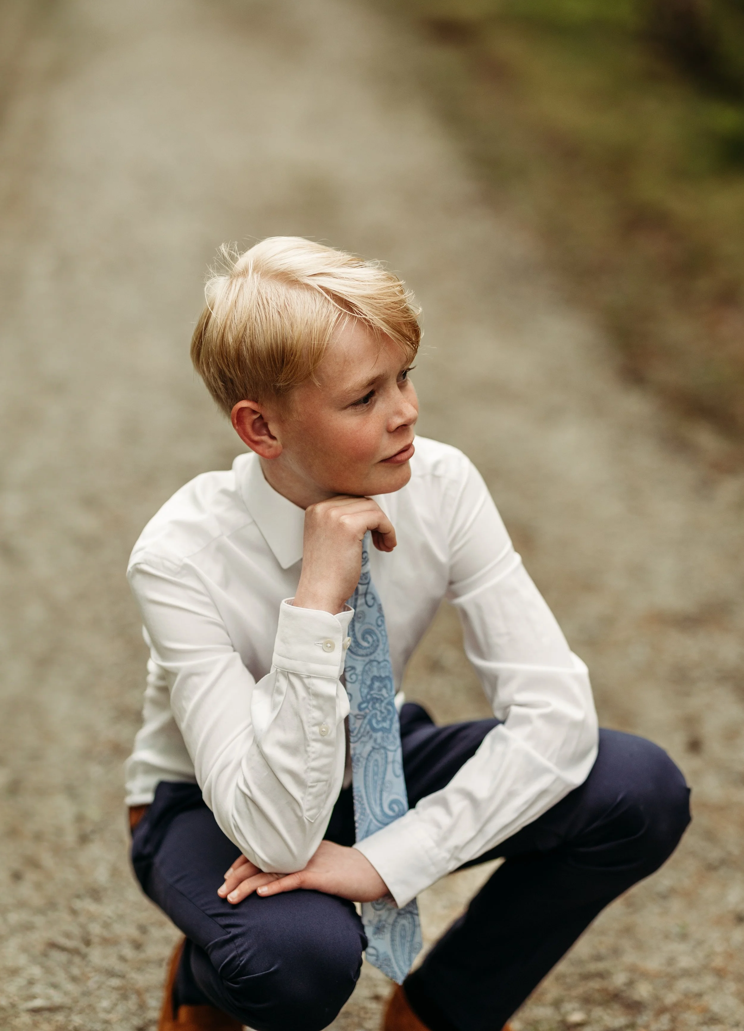 En ung gutt i dress som sitter på en vei, med hånd under haken, ser tankefull ut.