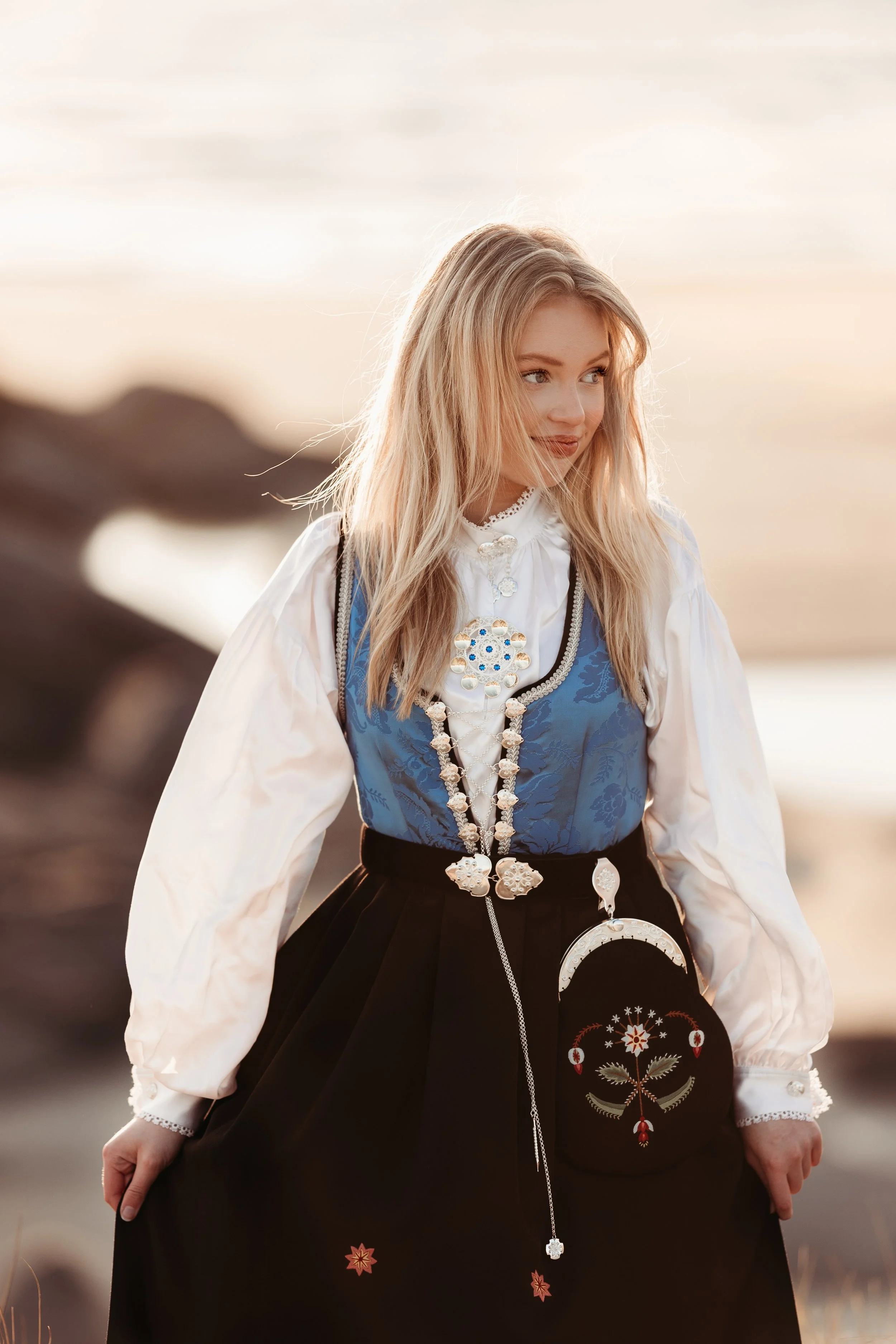 En ung kvinne i tradisjonell norsk bunad står utendørs ved solnedgang, med fjell og sjø i bakgrunnen.