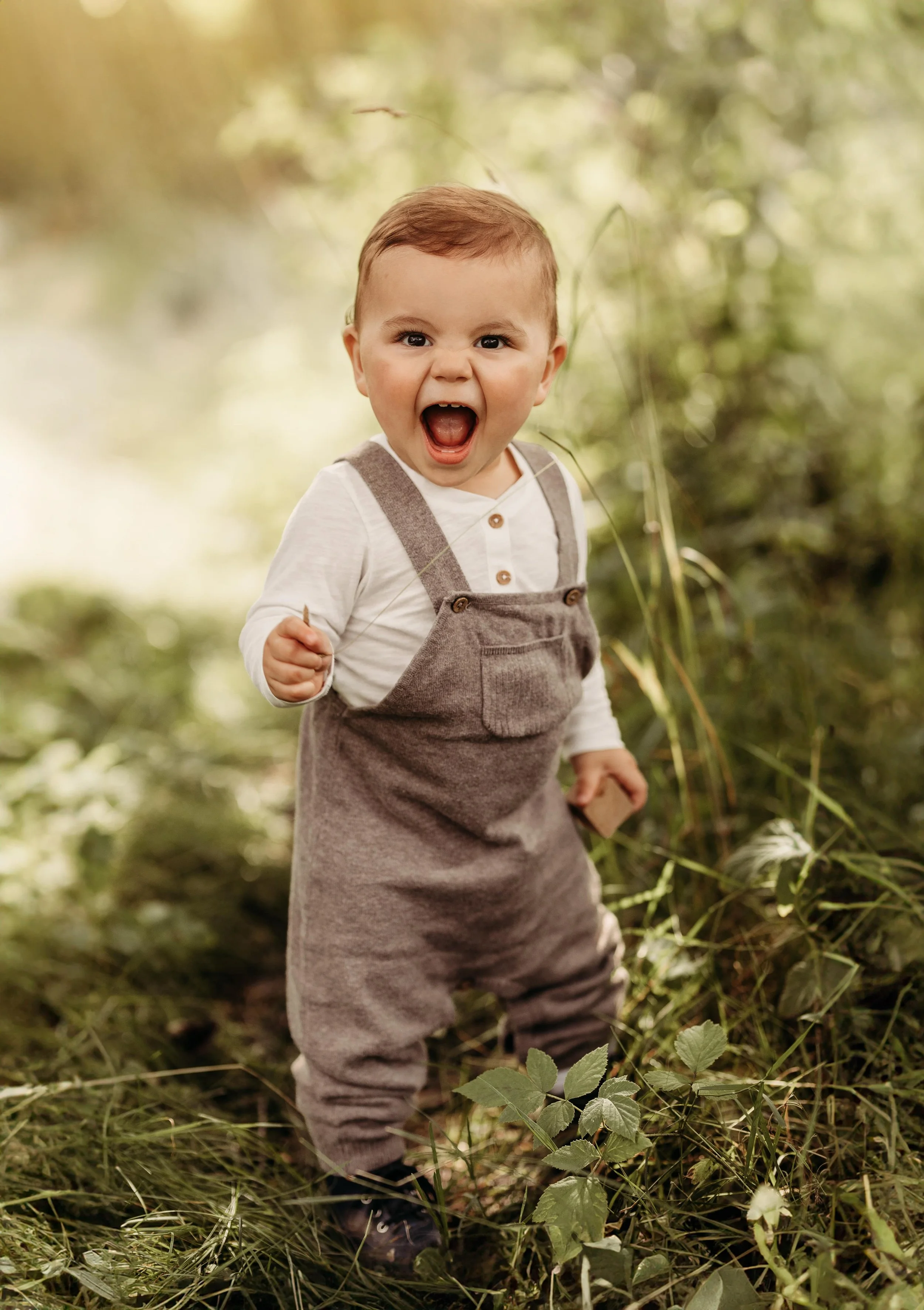 En liten gutt som står i en skog, smilende med åpen munn, iført grå bukser og en hvit skjorte.