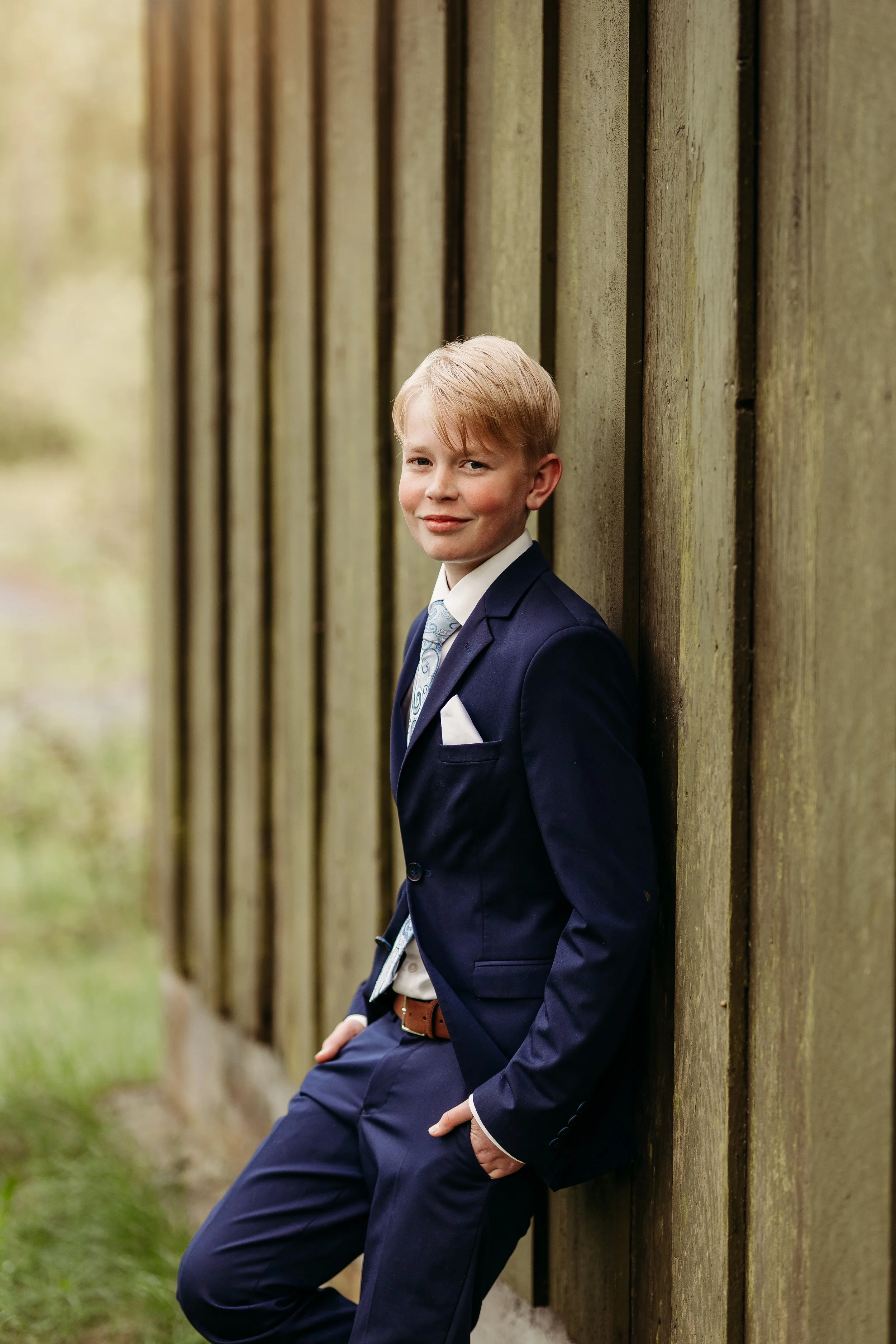 Ung mann i dress står ved en trevegg, utendørs.