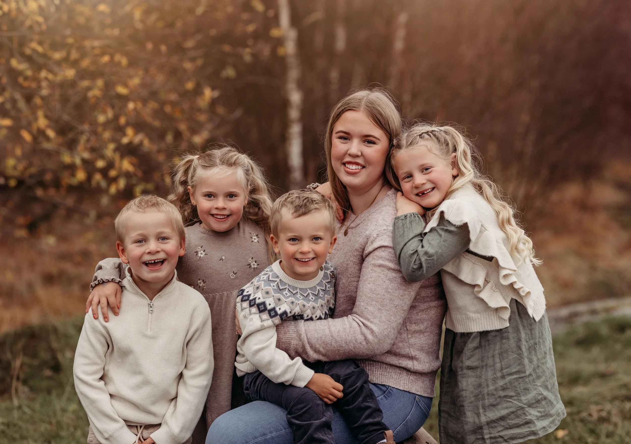 En kvinne med fire barn som smiler og klemmer hverandre utendørs i høstlige omgivelser.